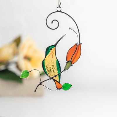 Stained glass hummingbird in green and yellow sitting on a branch with orange flower and green leaves