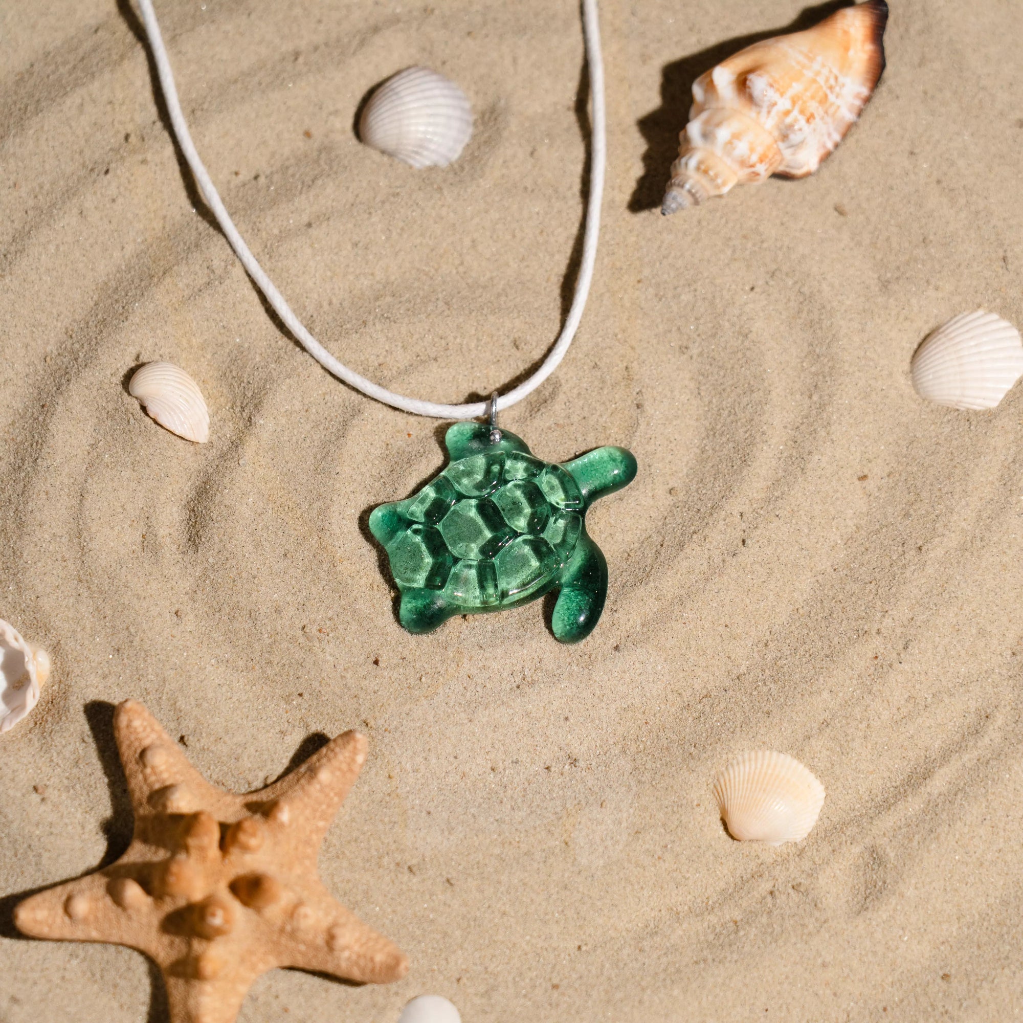 Emerald stained glass turtle pendant on a white cord with seashells and a starfish on sand