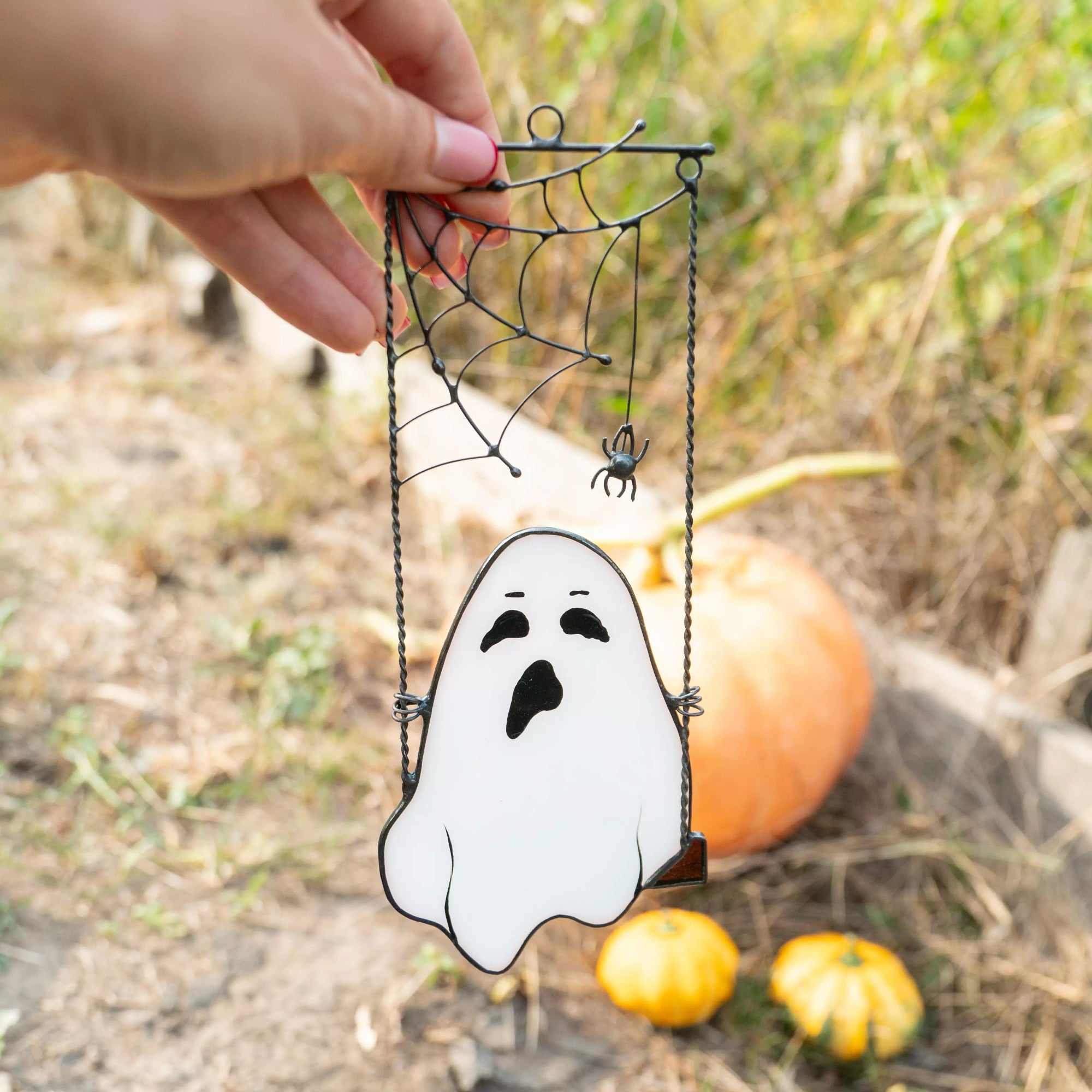 Hand holding ghost stained glass suncatcher with spiderweb near pumpkin garden