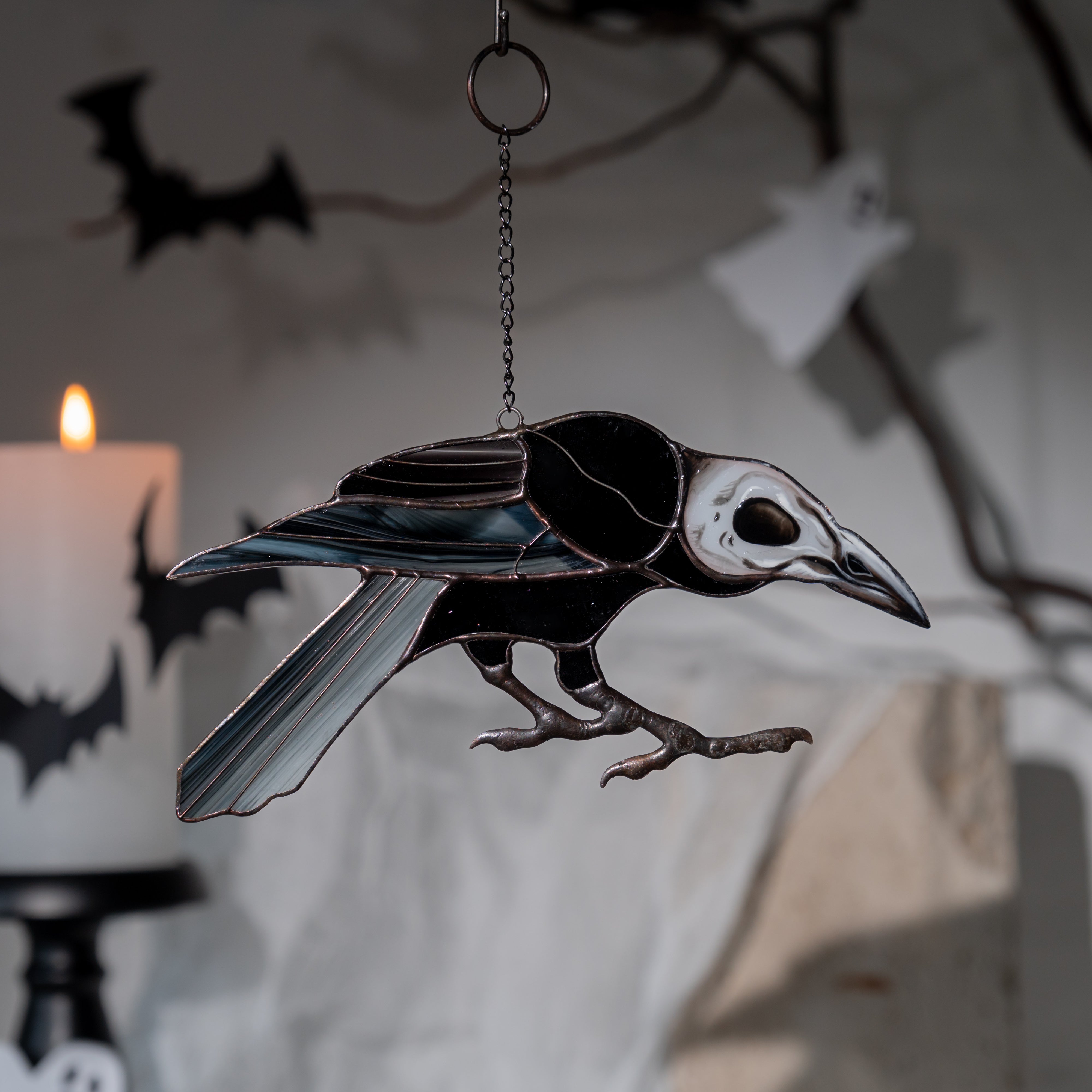 Raven suncatcher with skull head hanging in front of a candle and Halloween paper bats on a spooky indoor setup.
