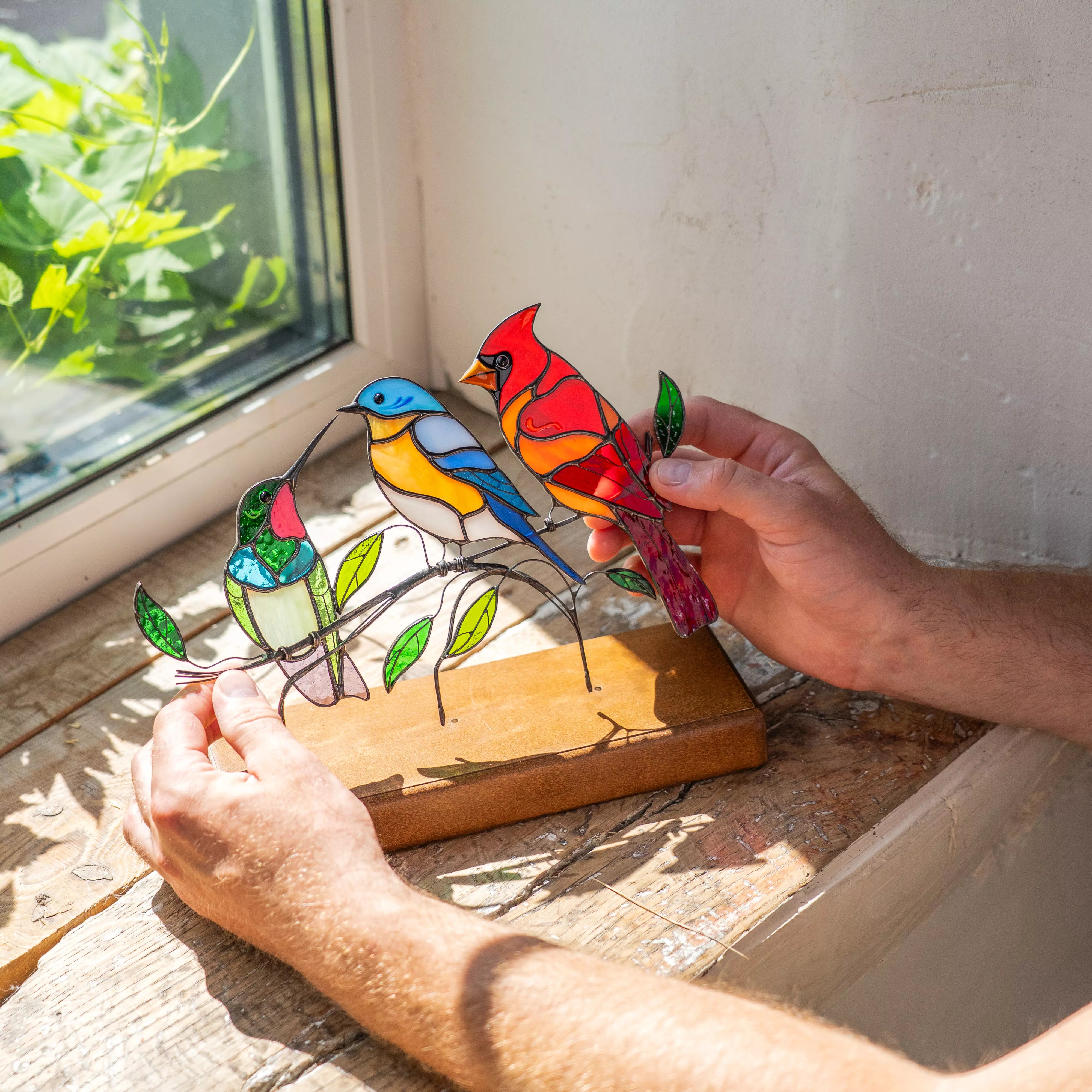 Hand holding a colorful stained glass birds on the wooden base by a window