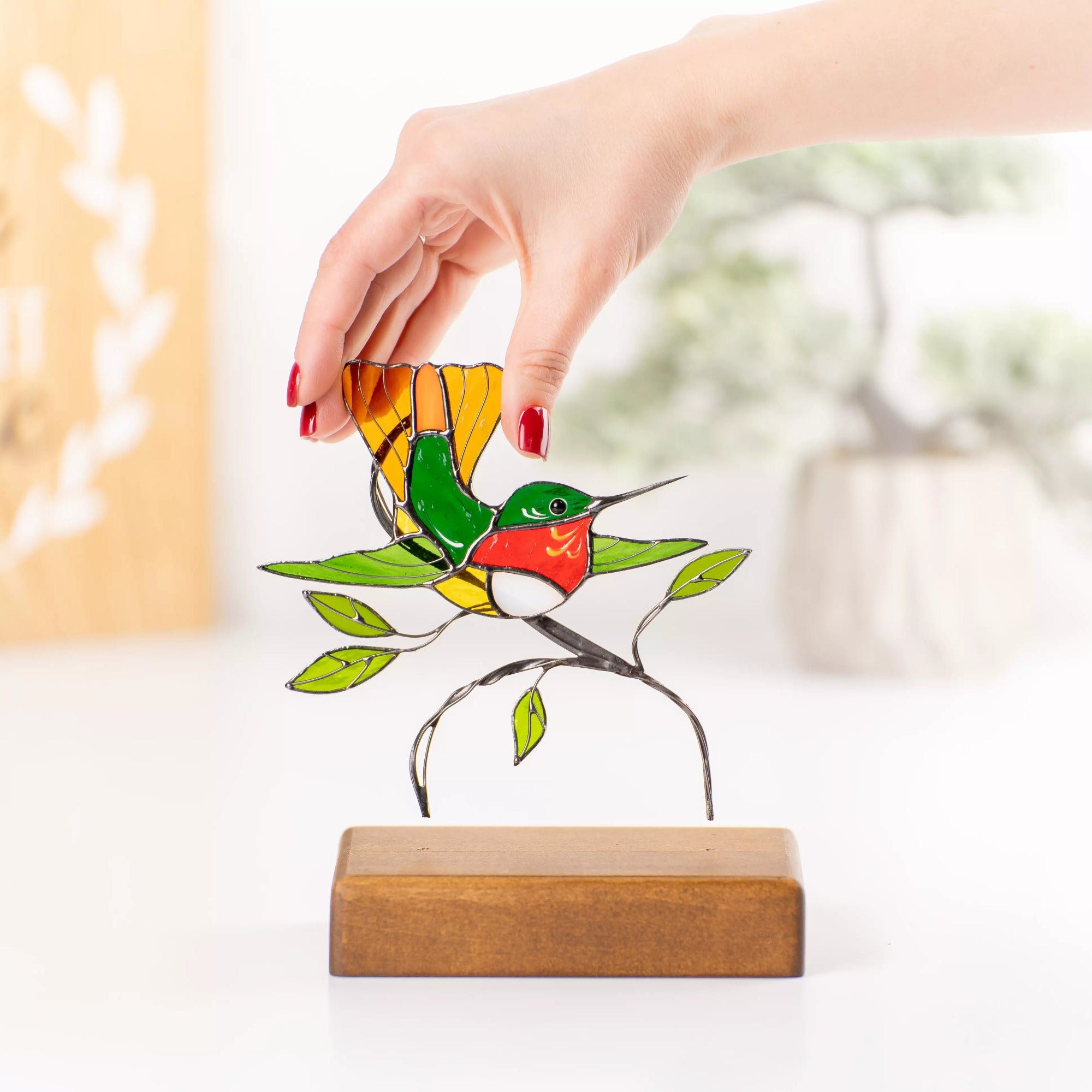 Hand holding a colorful stained glass hummingbird suncatcher near a wooden base