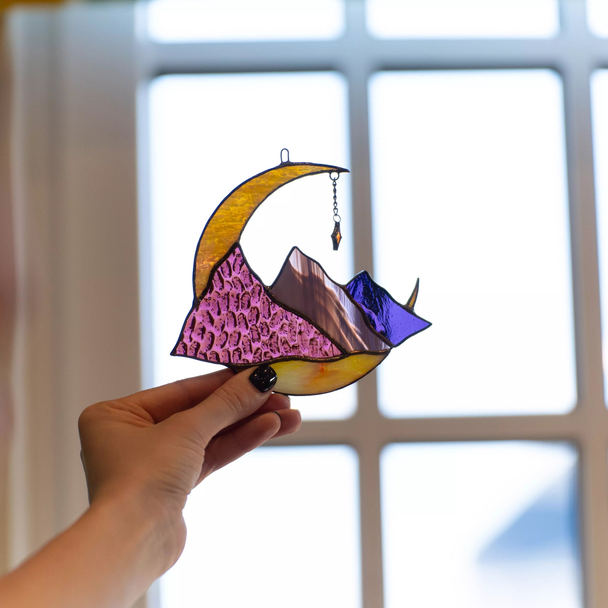 Hand holding a vibrant stained glass moon and mountain suncatcher in front of a window.