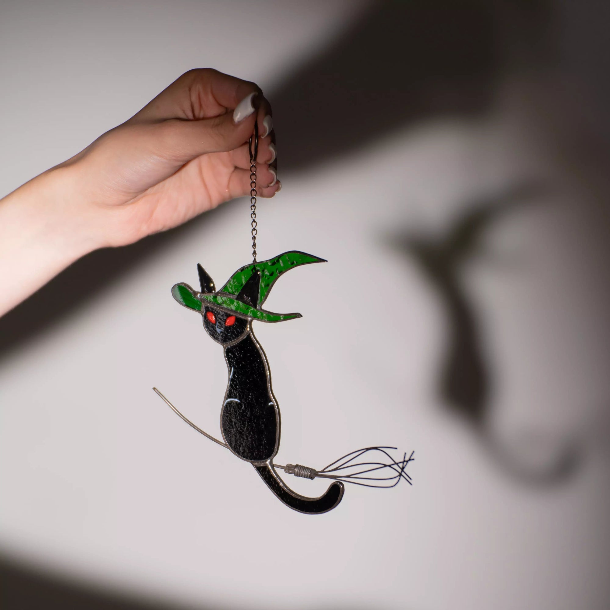 Hand holding a stained glass black cat with green hat and red eyes, casting a shadow on a wall
