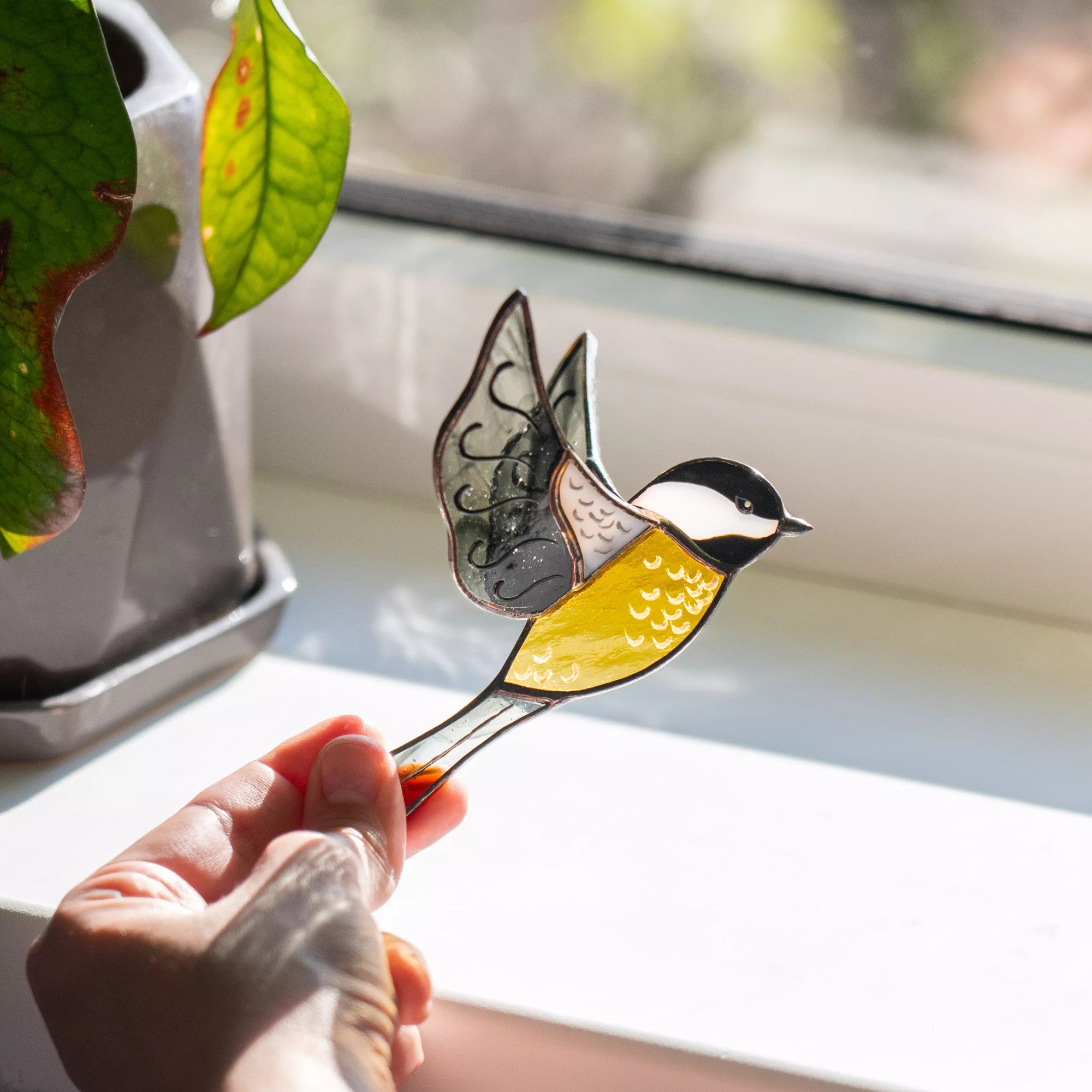 Hand holding a stained glass chickadee bird with a butterfly on a windowsill.