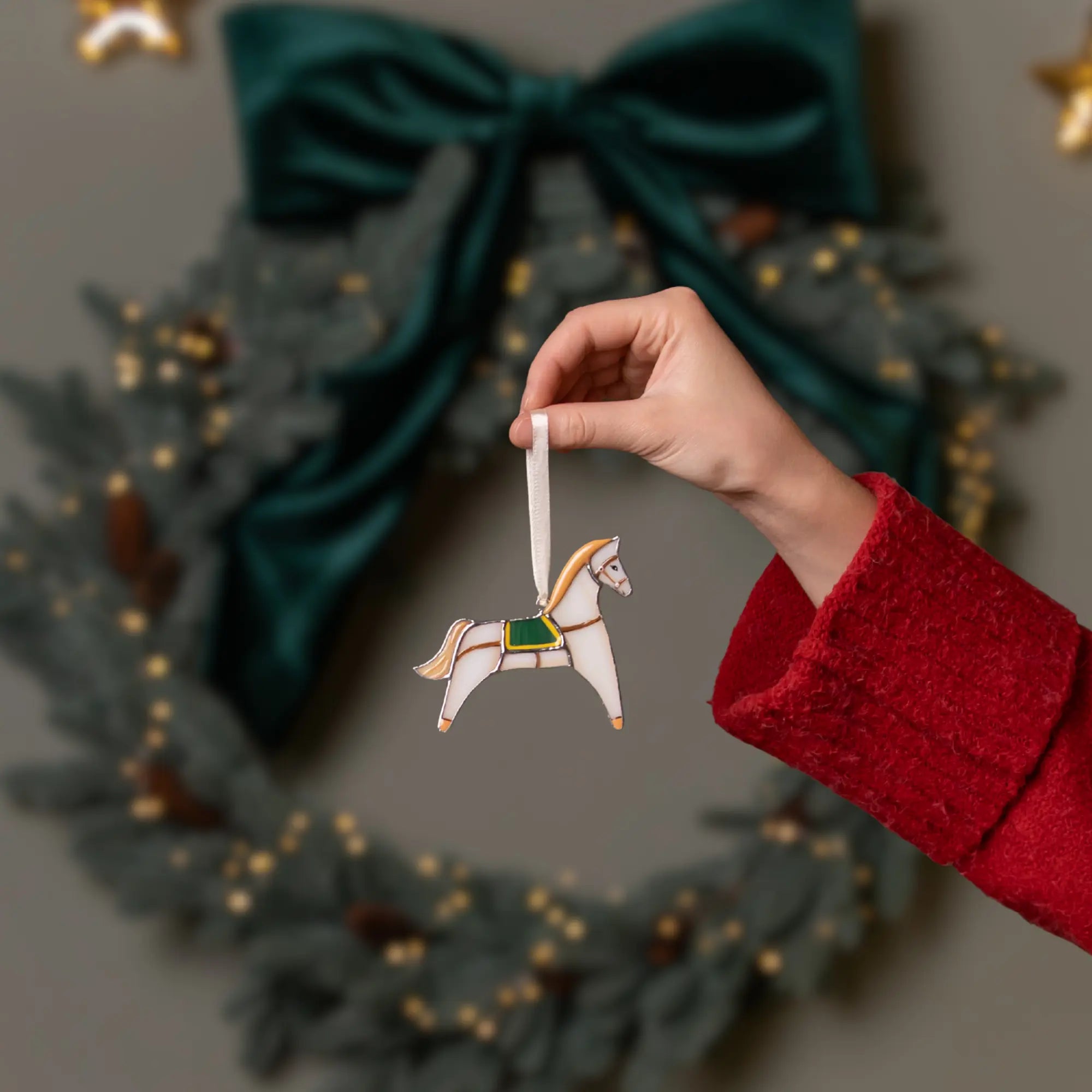 Hand holding a stained glass horse ornament in front of a Christmas wreath with a green bow.