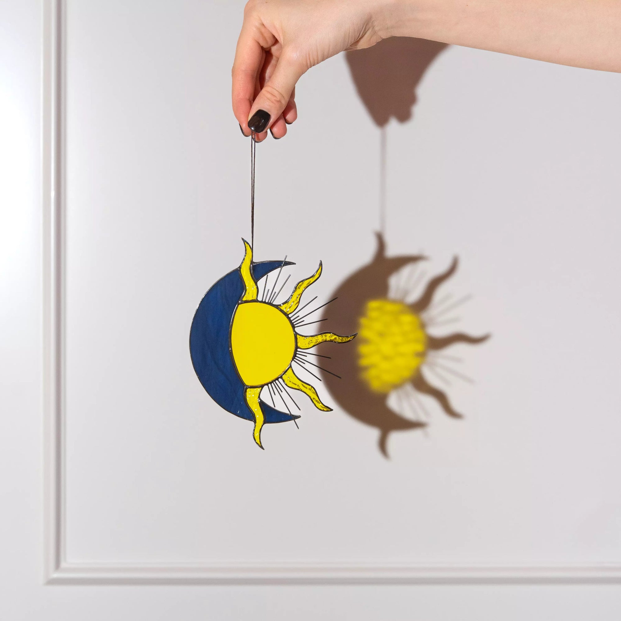 Hand holding a decorative sun and moon ornament casting a shadow on a light gray background