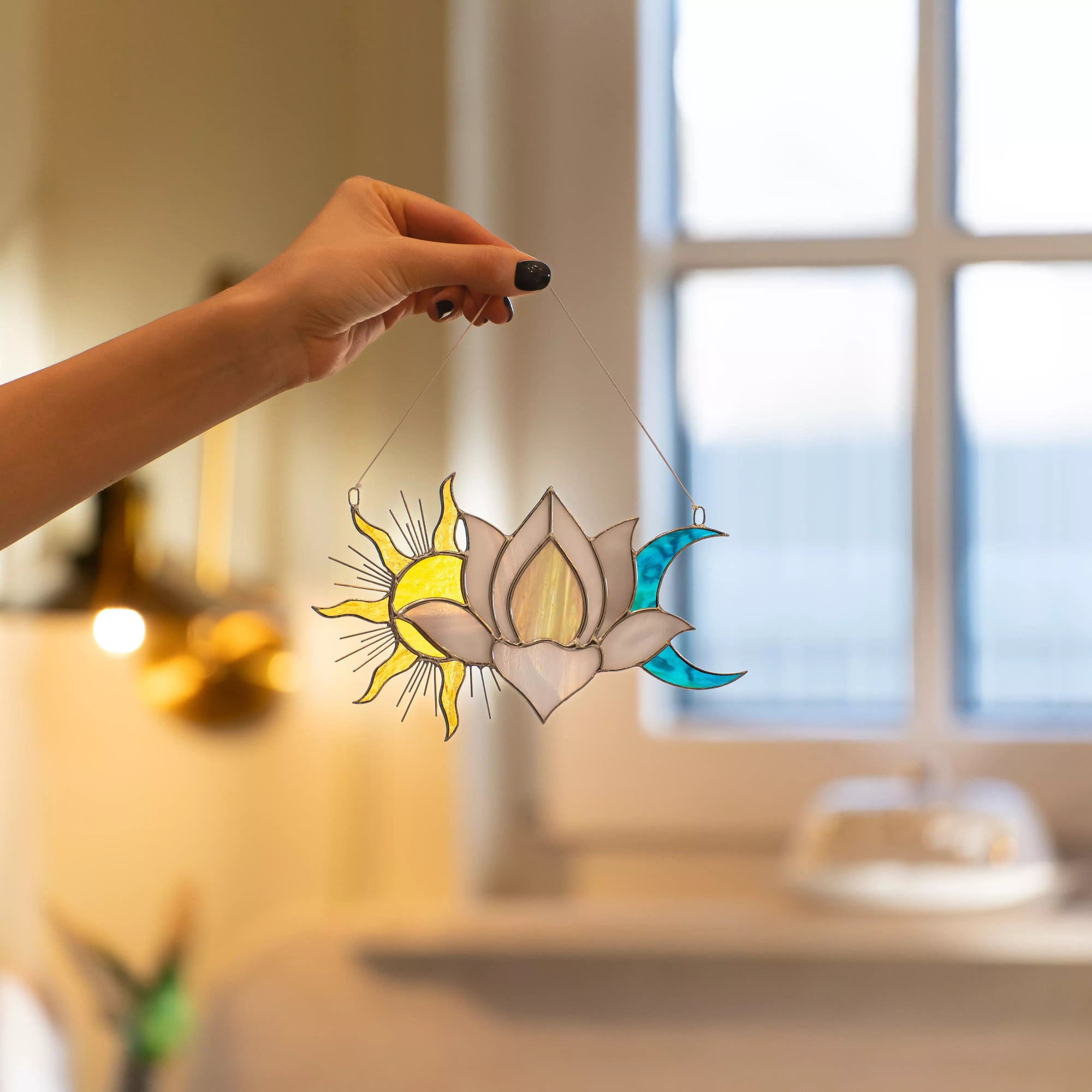 Hand holding a decorative sun, moon, and lotus flower window hanging in front of a window.