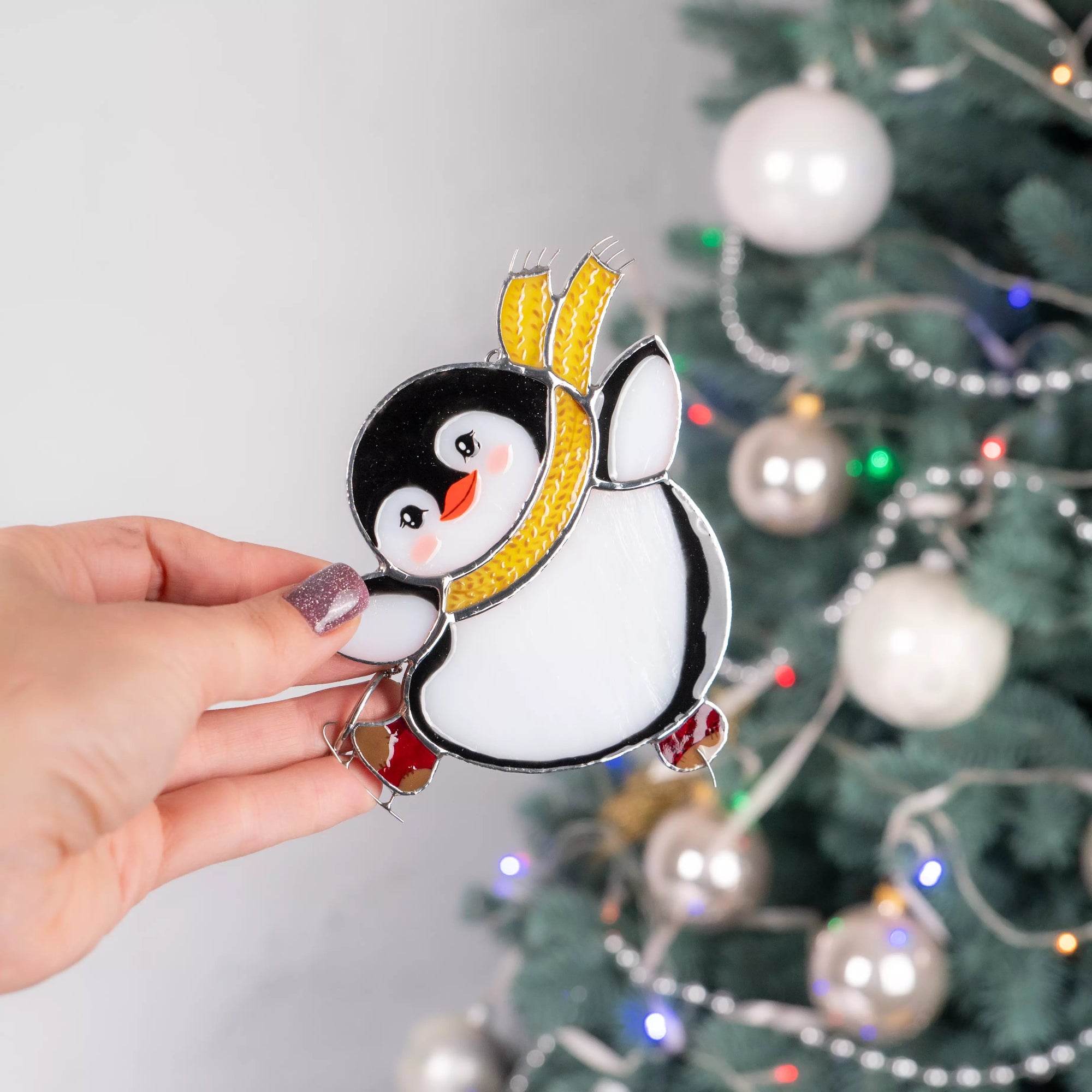 Hand holding a penguin stained glass ornament with a Christmas tree in the background