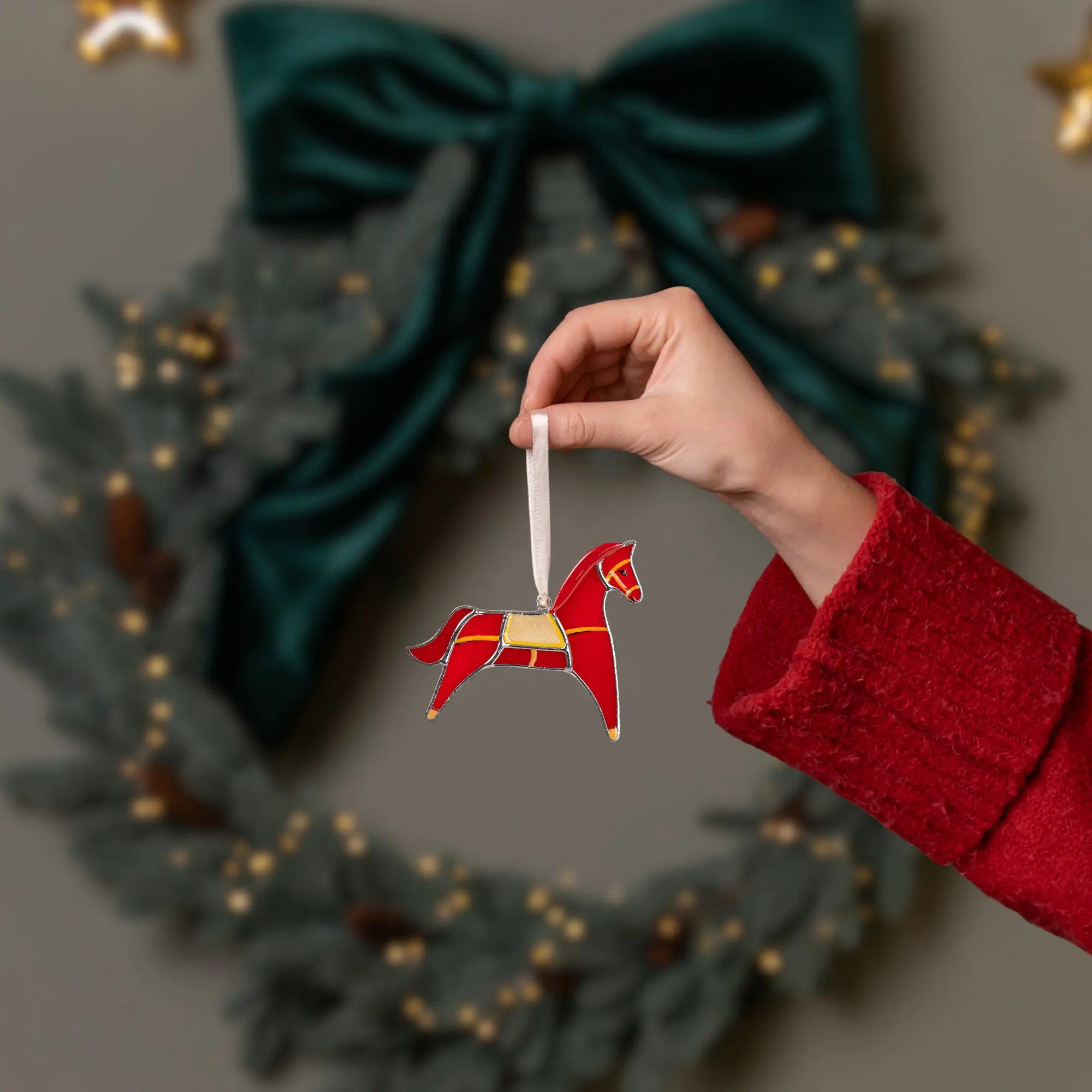 Hand holding a red horse stained glass ornament in front of a Christmas wreath.