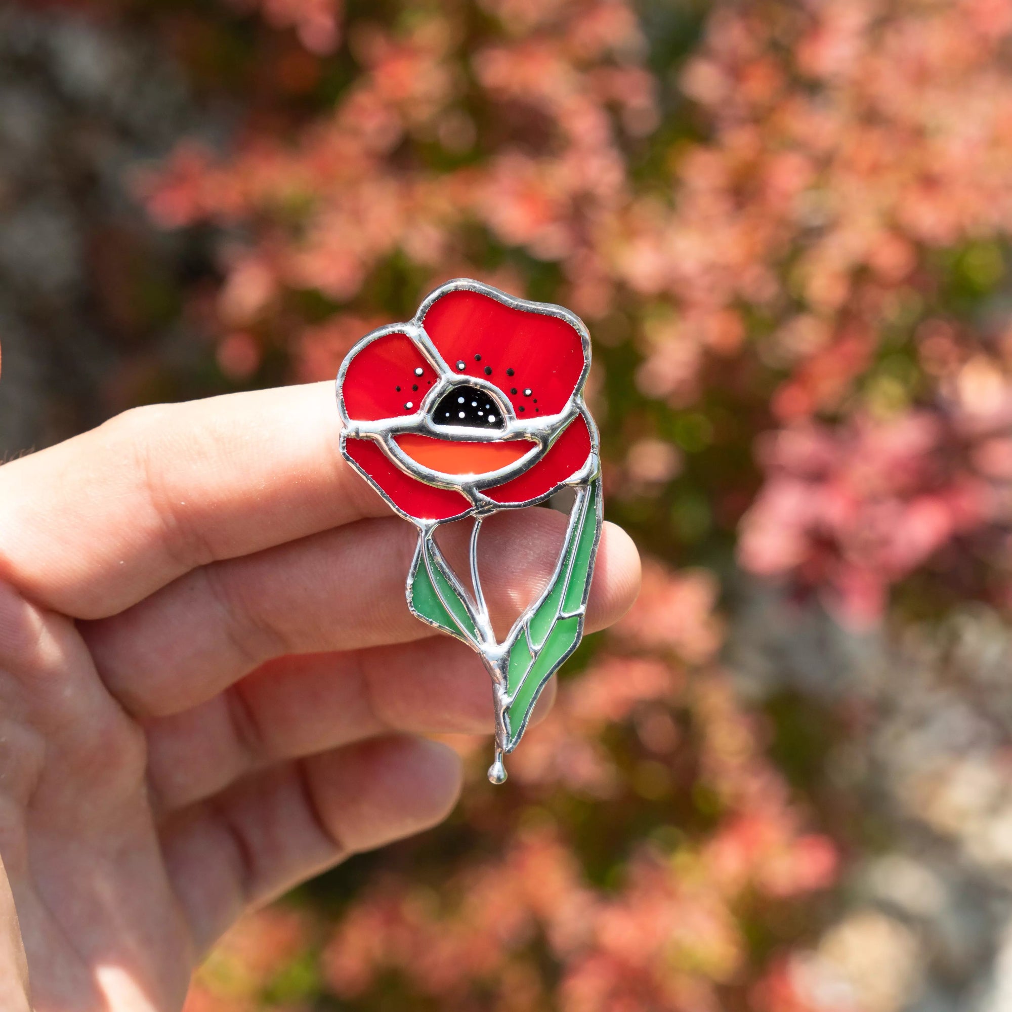 Hand holding stained glass red poppy stained glass pin with a blurred natural background