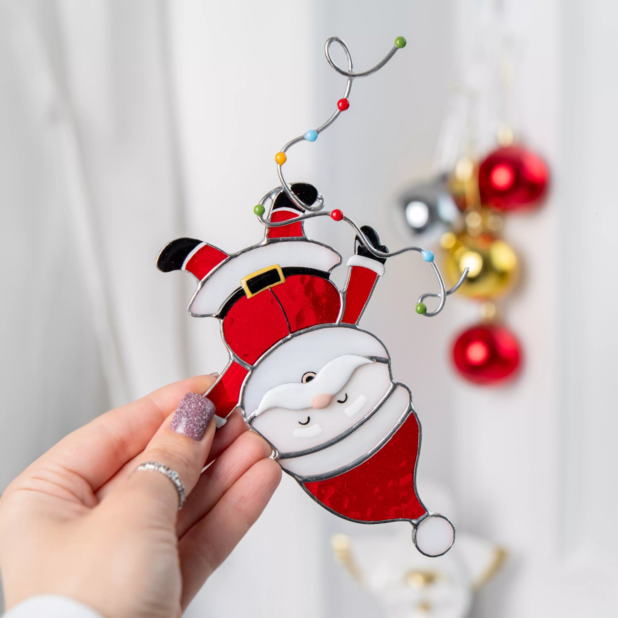 Hand holding a stained glass Santa Claus suncatcher with a blurred Christmas tree in the background.
