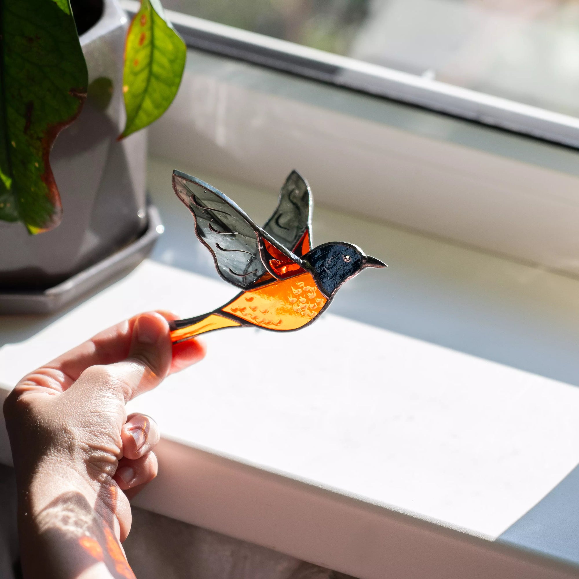 Hand holding a stained glass baltimore oriole decoration by a window