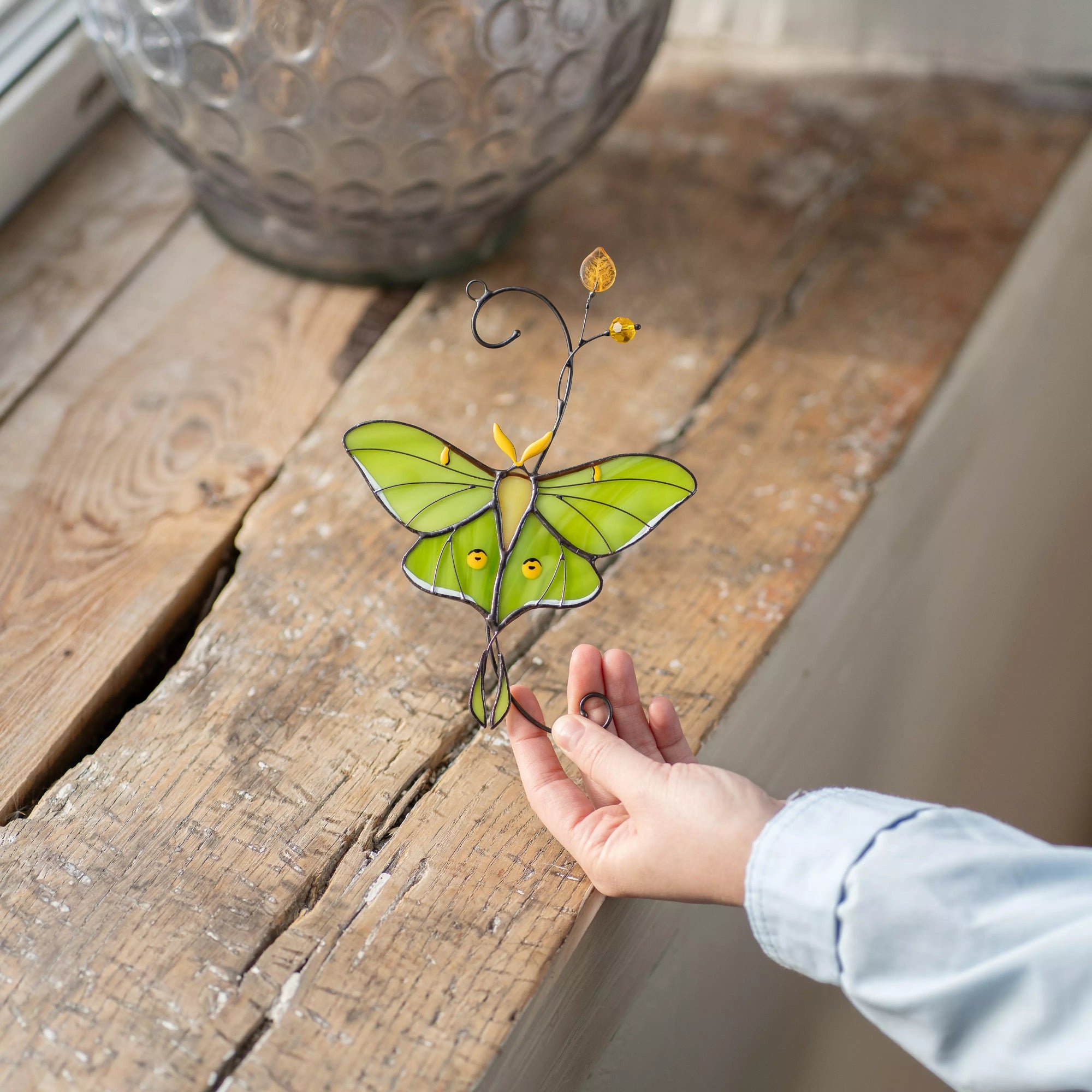 Hand holding a stained glass luna moth butterfly suncatcher on a wooden surface