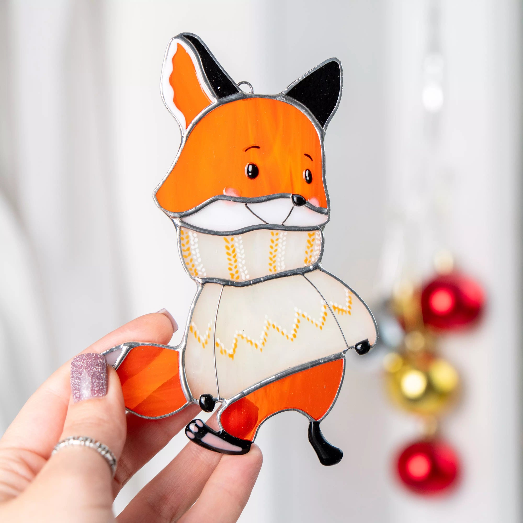 Hand holding a stained glass suncatcher fox ornament with a Christmas decorations in the background