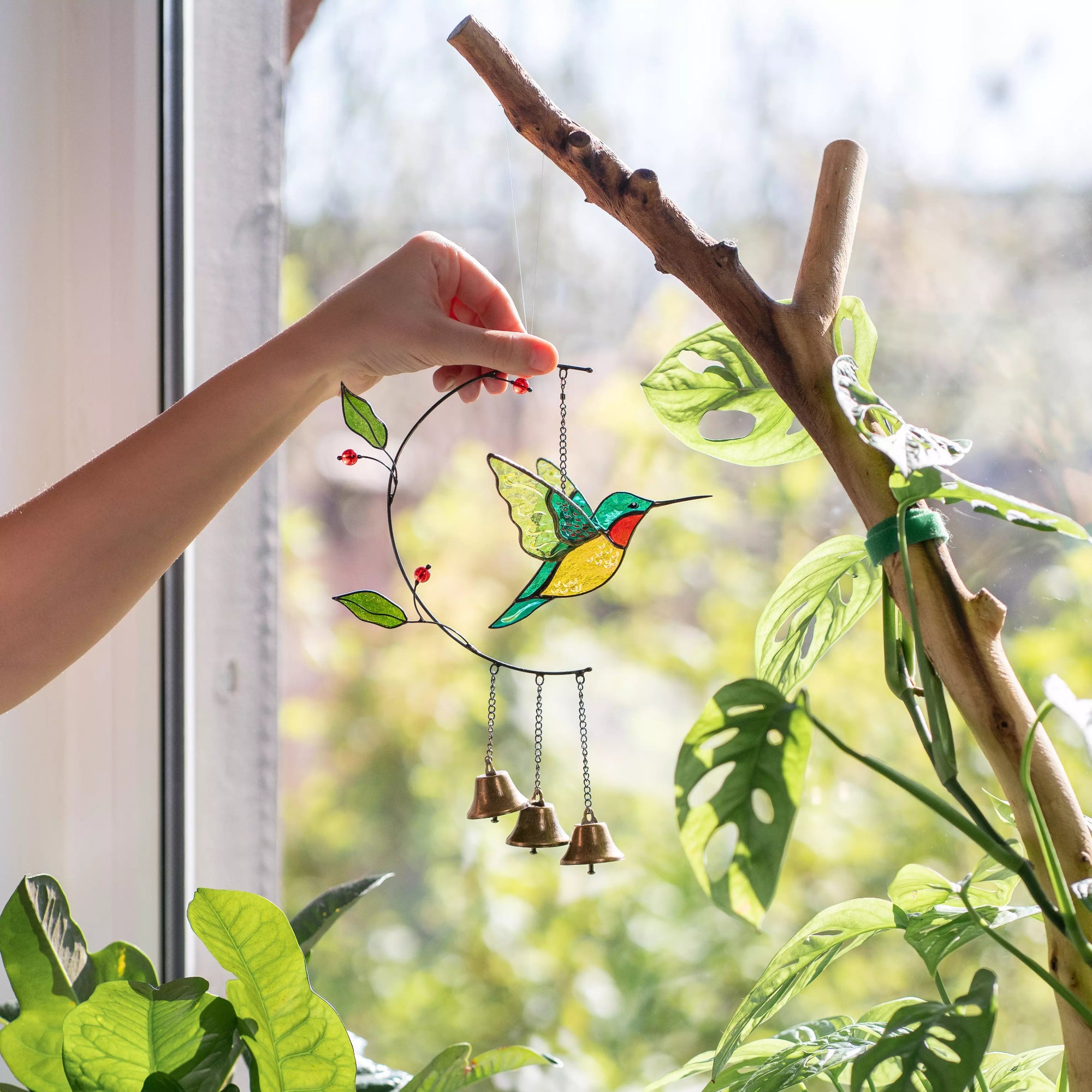 Hand holding a stained glass hummingbird wind chime with bells against a window with greenery outside.