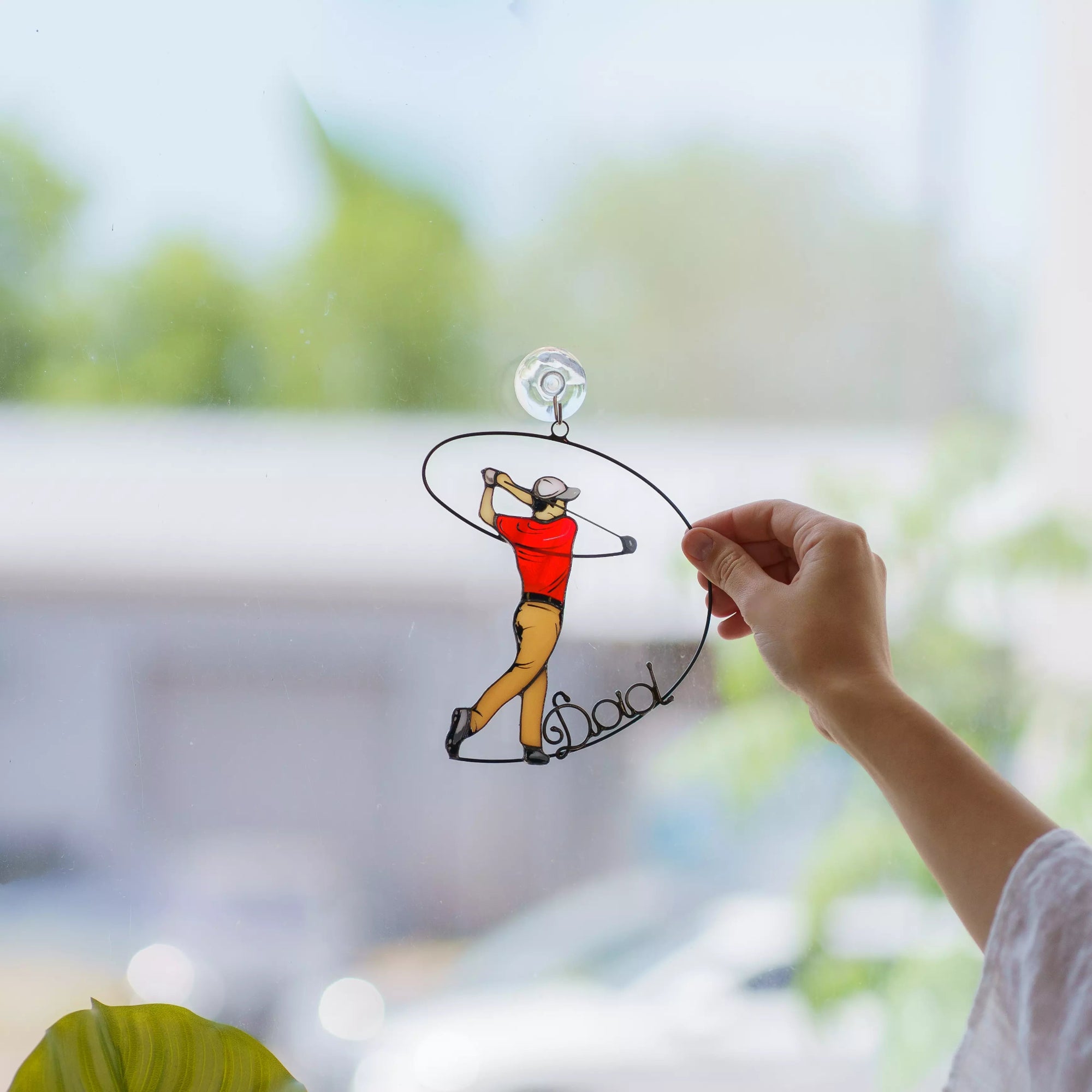 Hand holding a stained glass suncatcher of a golfer with 'Dad' on a blurred green outdoor background