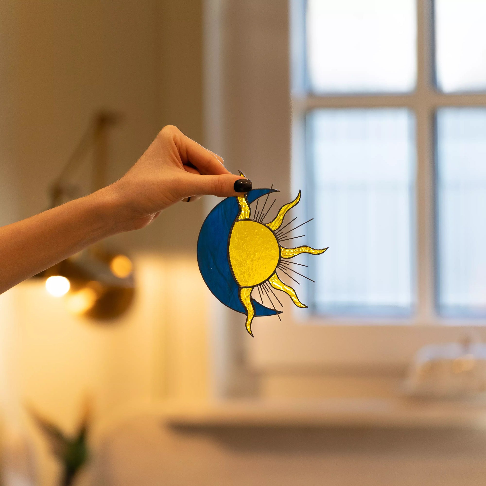 Hand holding a sun and moon stained glass suncatcher in an indoor setting
