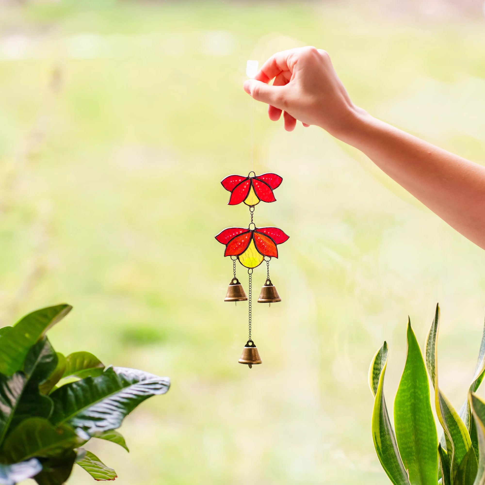 A hand holding a vibrant red and yellow stained glass flower wind chime against a bright, softly blurred green outdoor background.