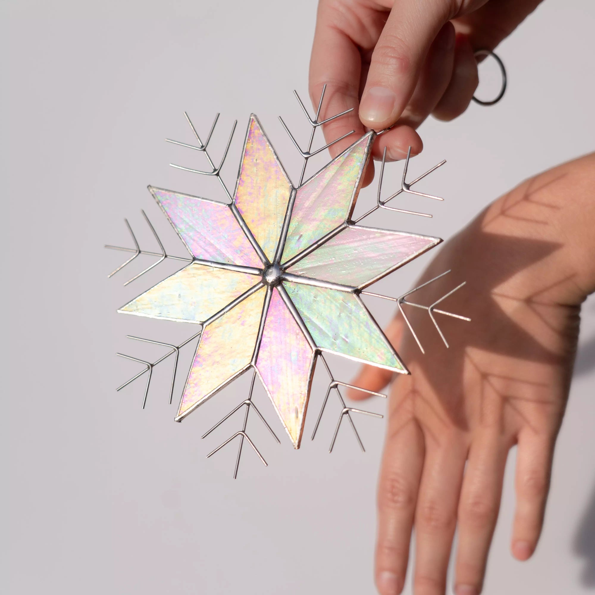 Hand holding an iridescent snowflake stained glass ornament, casting a soft shadow on the other hand, against a light background