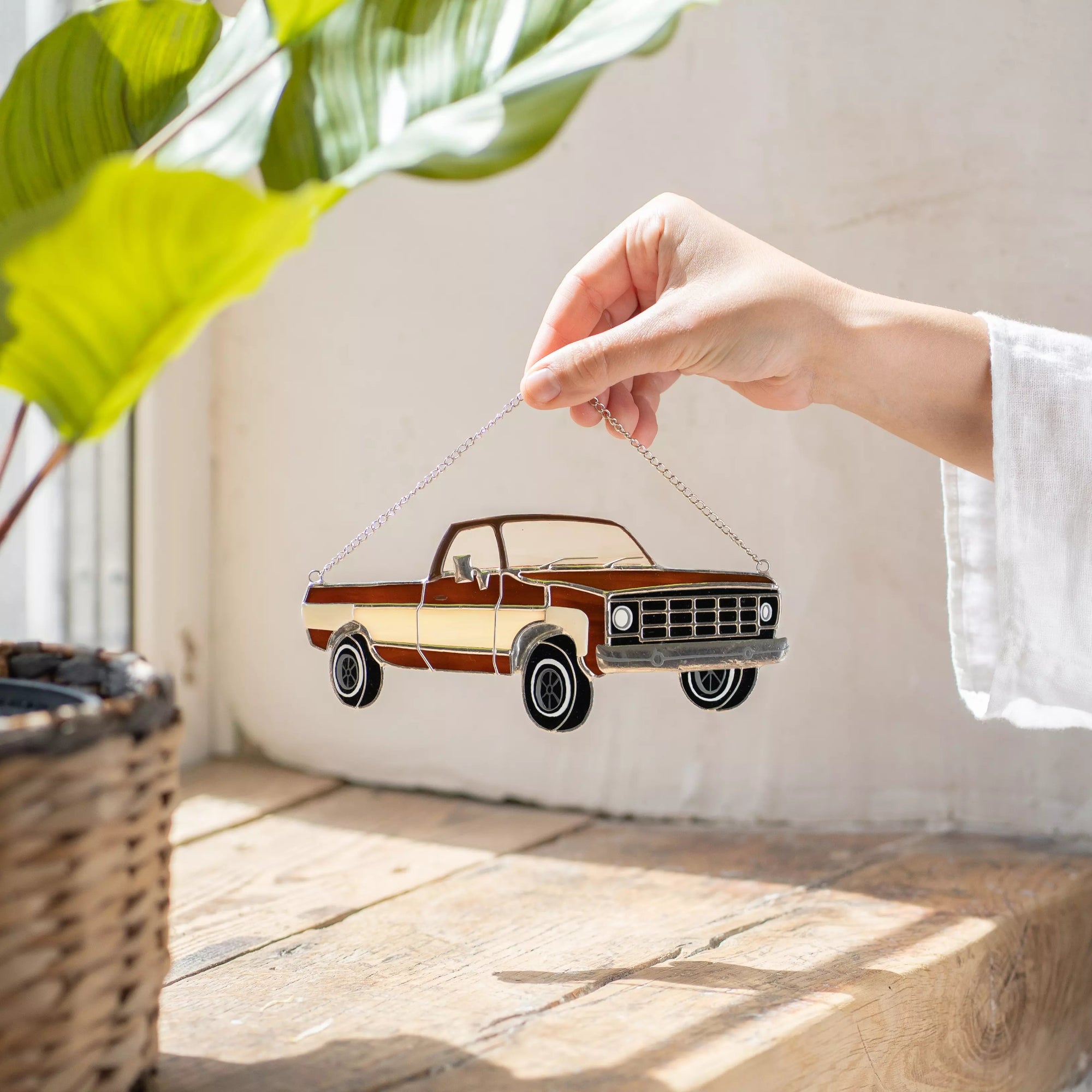 Hand holding stained glass model of a vintage truck, with a plant and woven basket in the background.