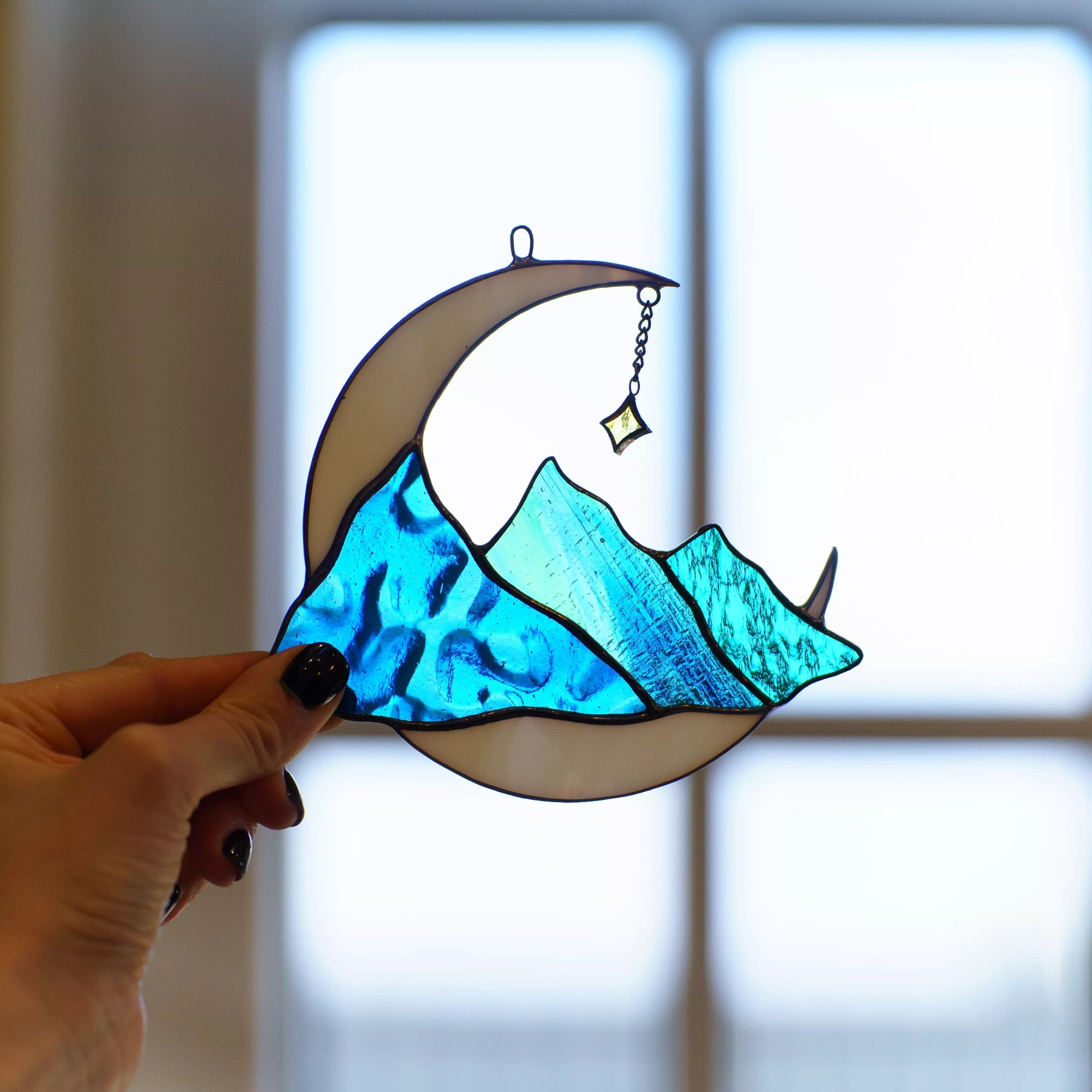 Hand holding stained glass celestial suncatcher moon with blue mountains in front of a window