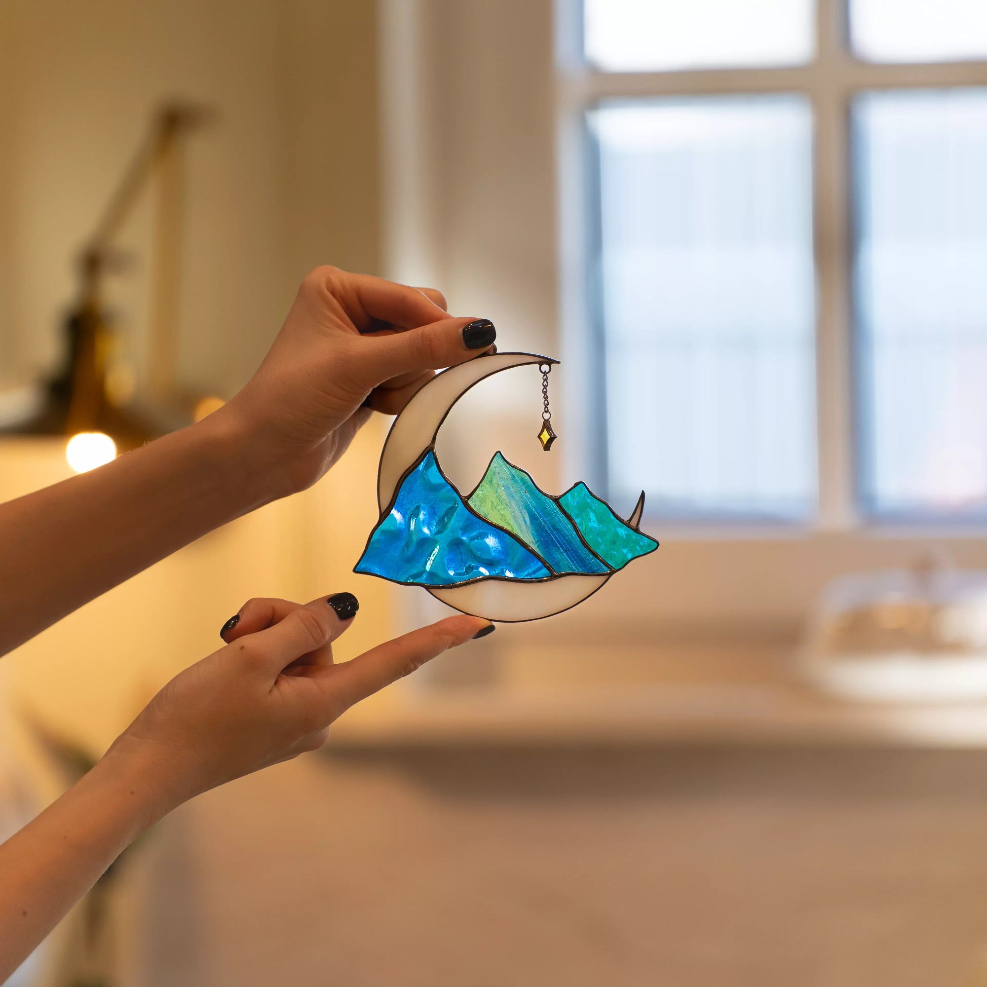 Hand holding stained glass celestial suncatcher moon with blue mountains in a warm cozy interior