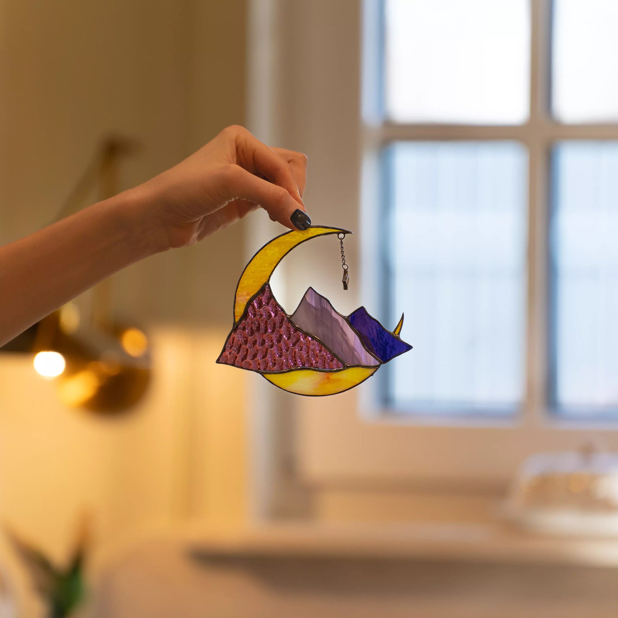 Hand holding vibrant stained glass crescent moon and mountain suncatcher in cozy home interior