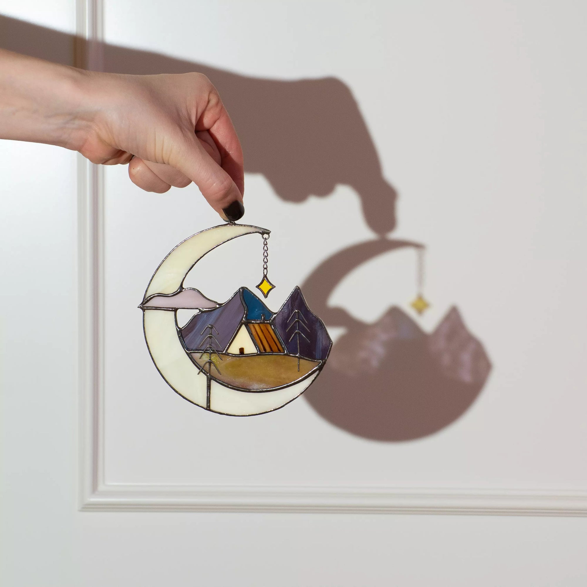 Hand holding stained glass suncatcher with a crescent moon, mountain landscape, and house.
