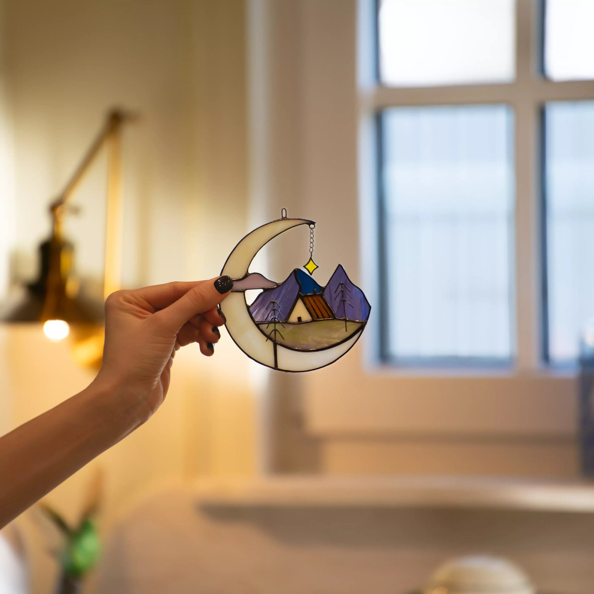 Hand holding stained glass suncatcher featuring a crescent moon, mountain landscape, and house in a cozy interior
