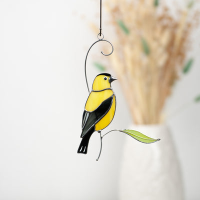 Yellow and black stained glass goldfinch perched with a green glass leaf, perfect for window decor.