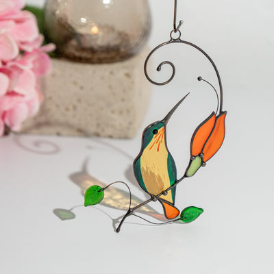 Green and yellow stained glass hummingbird near orange flower, casting colorful shadow on white surface