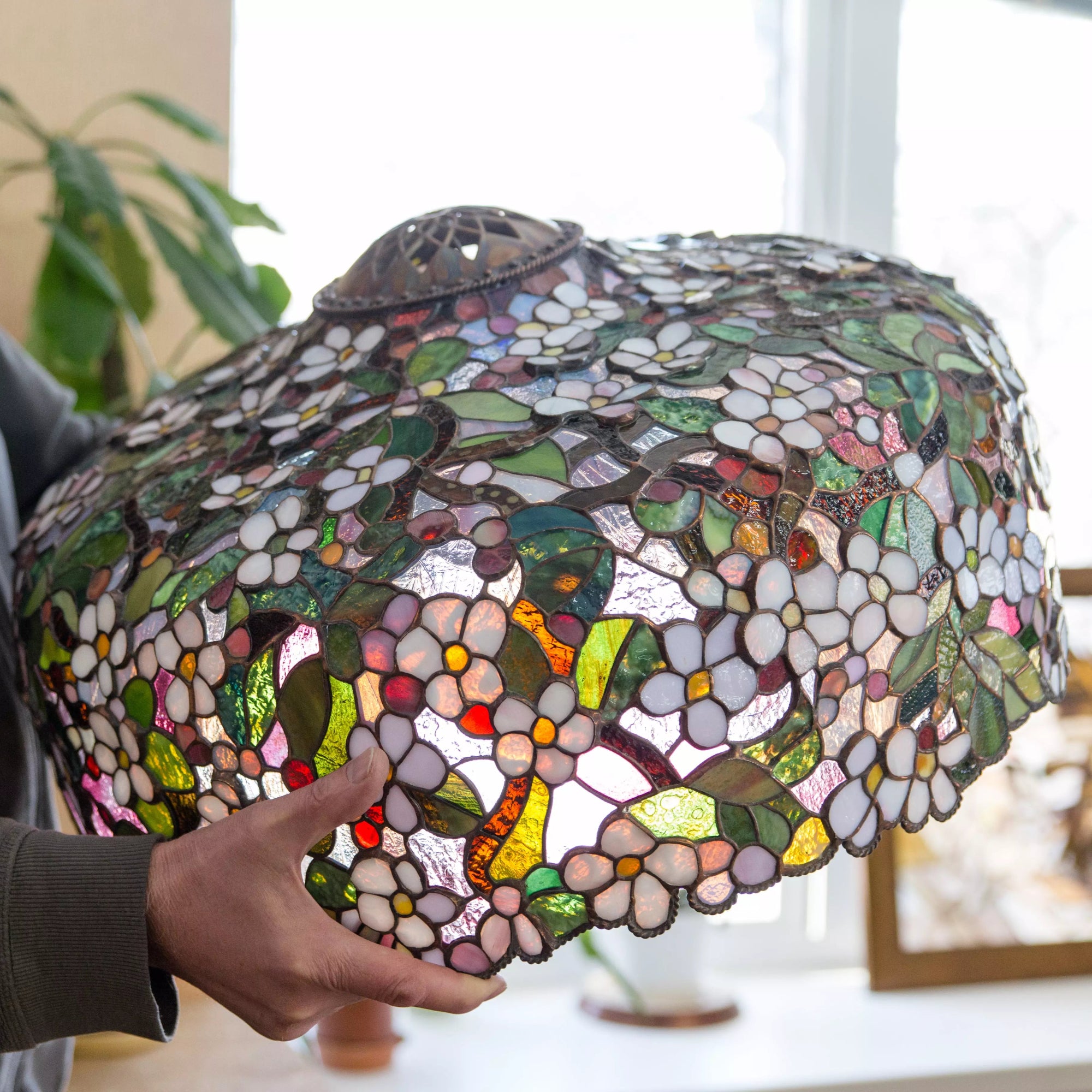 Person holding stained glass chandelier with colorful floral design in natural light near a window.