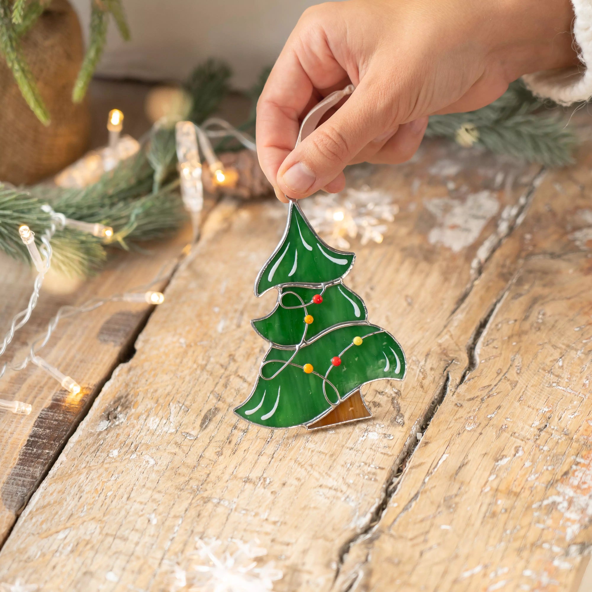 Green stained glass Christmas tree suncatcher with colorful garland placed on a rustic wooden table with fairy lights.
