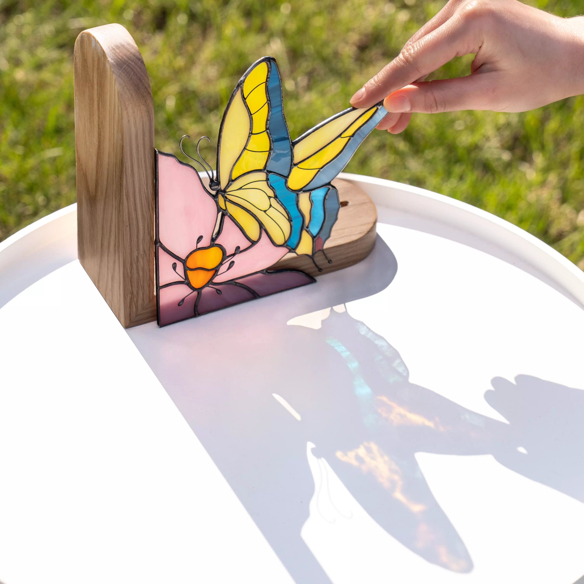 Hand touching a stained glass butterfly bookend placed in a wooden stand on a white table, showing scale, movable wing detail, and colorful glass reflections in sunlight.
