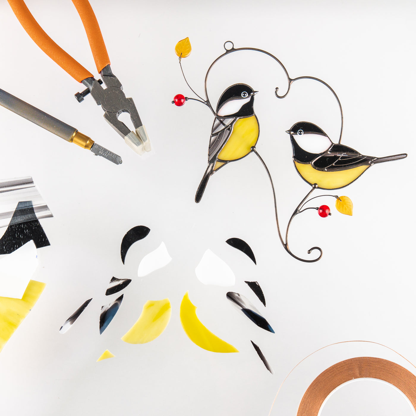 Two stained glass chickadees with yellow bellies and red beads shown alongside glass cutting tools and glass pieces.