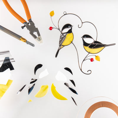 Two stained glass chickadees with yellow bellies and red beads shown alongside glass cutting tools and glass pieces.