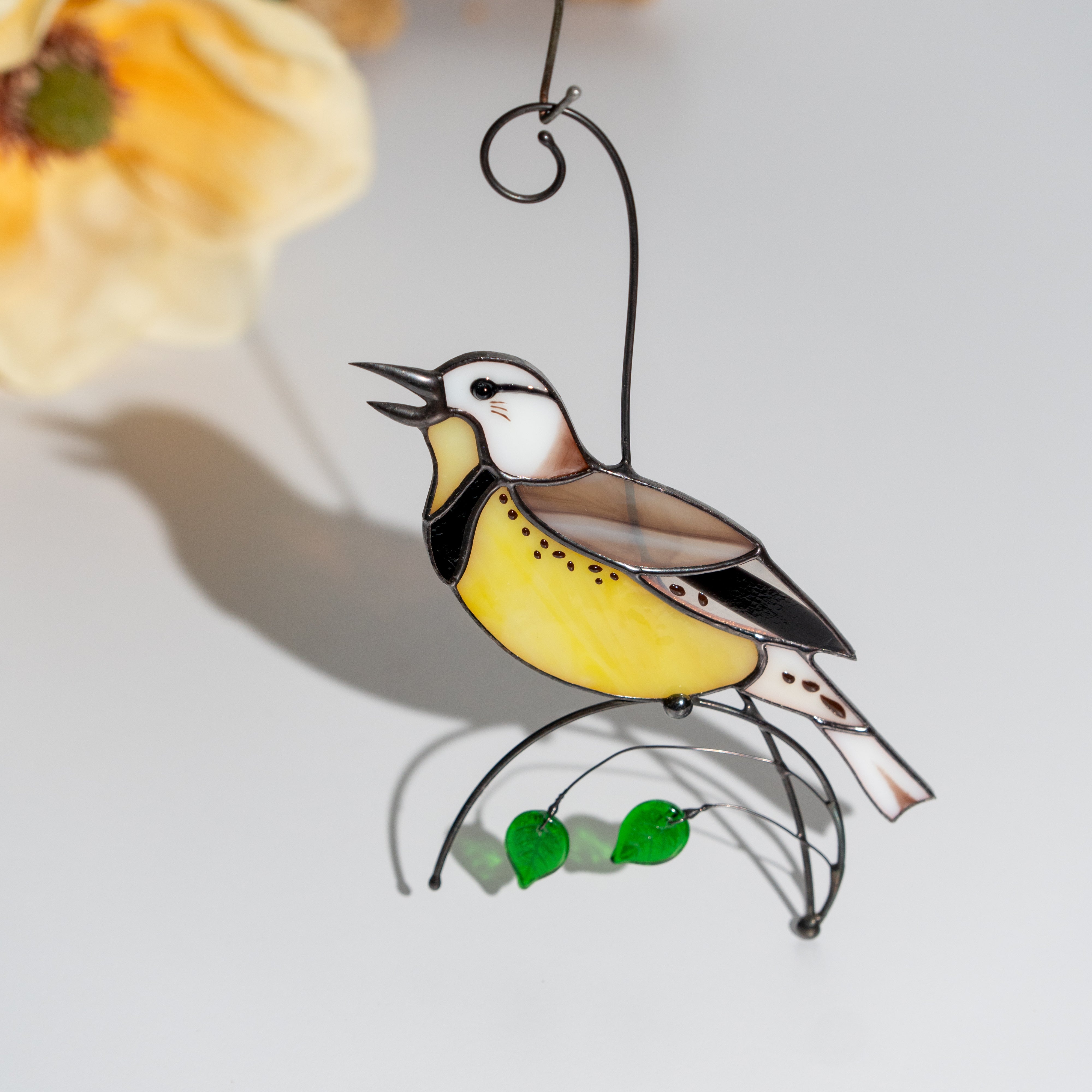 Yellow stained glass meadowlark bird with green glass leaves, placed near soft yellow flower, sunlight casting shadow