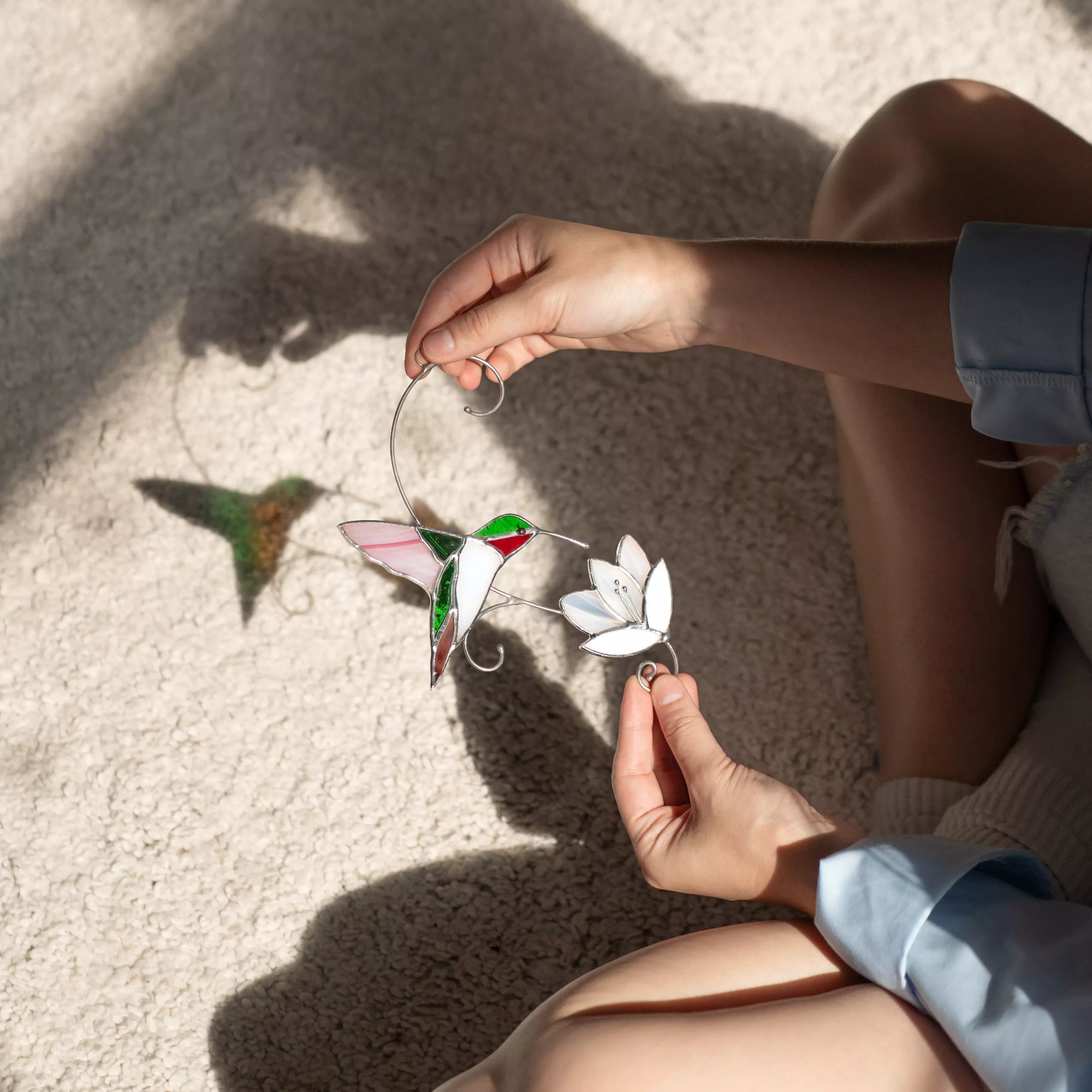 Stained glass hummingbird suncatcher in natural setting, shown with soft shadow on carpet as woman admires it.