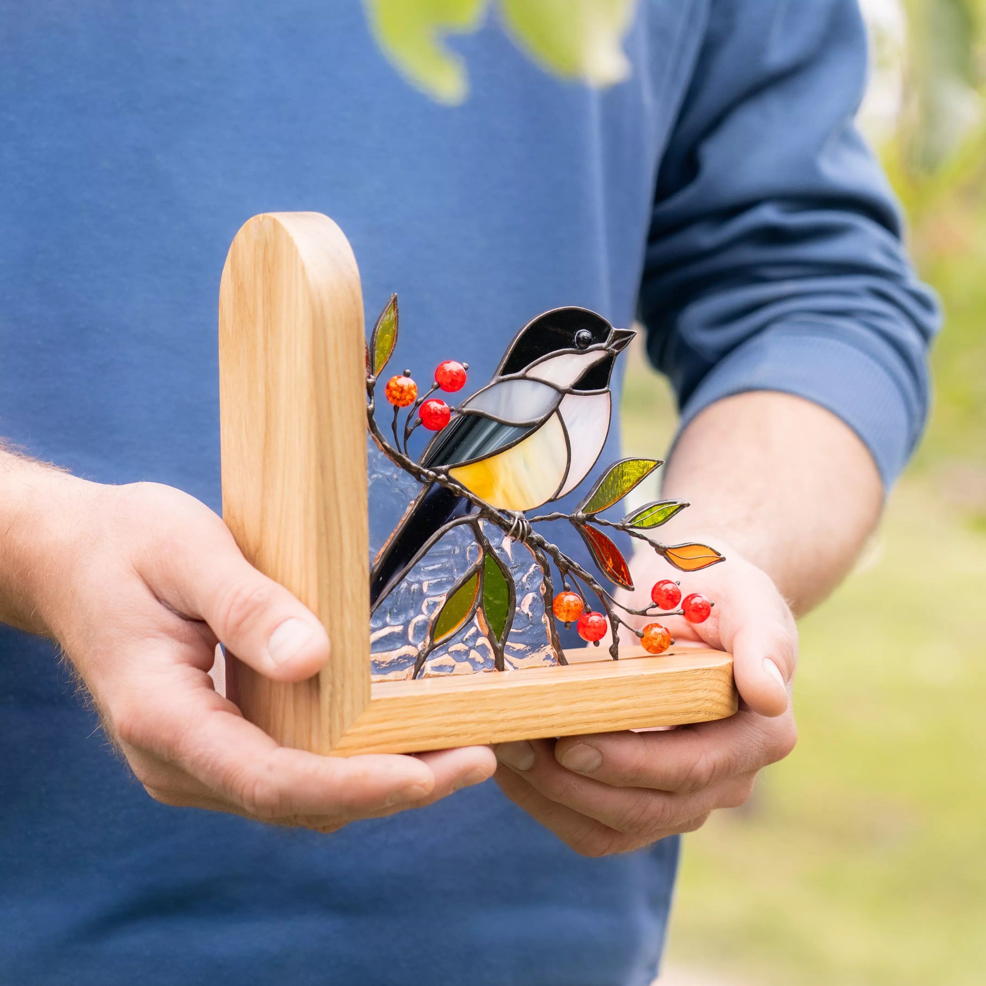Person holding a wooden frame with a stained glass bird design against a blurred natural background