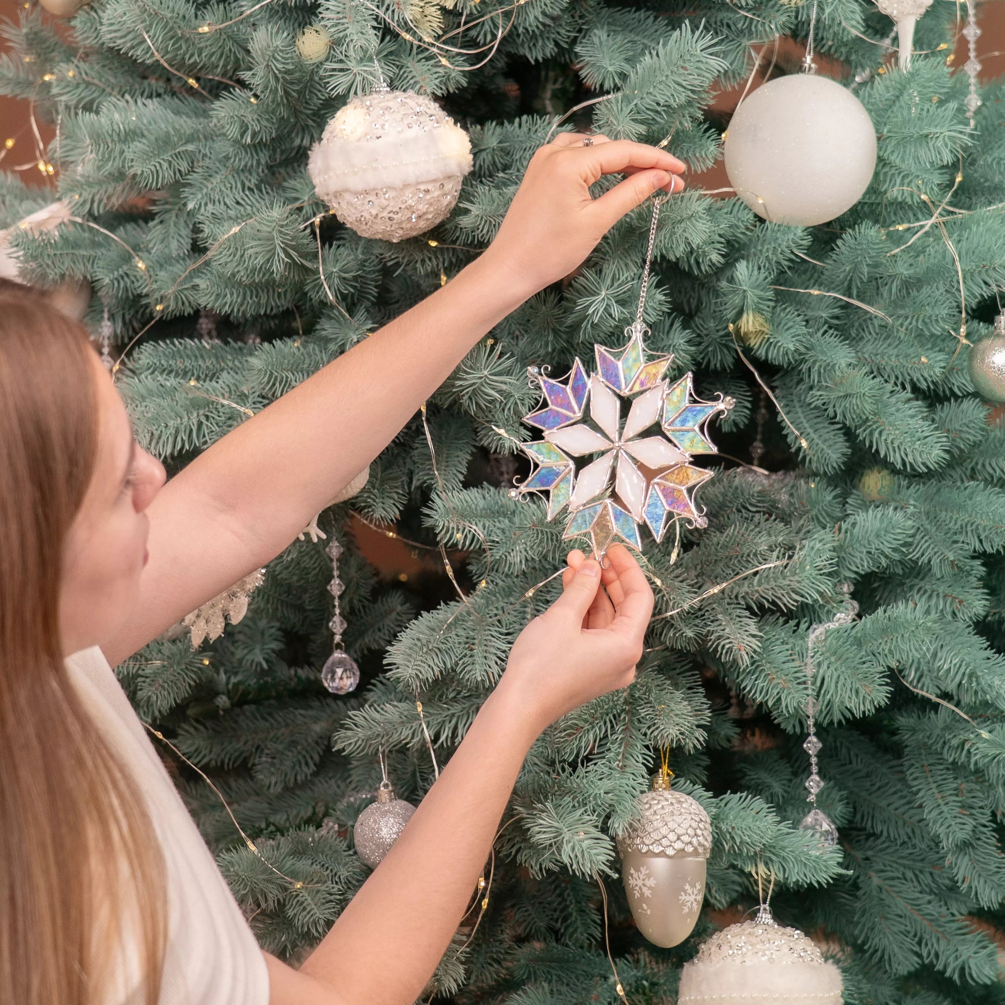 Woman decorating Christmas tree with a large iridescent stained glass snowflake ornament among silver baubles.