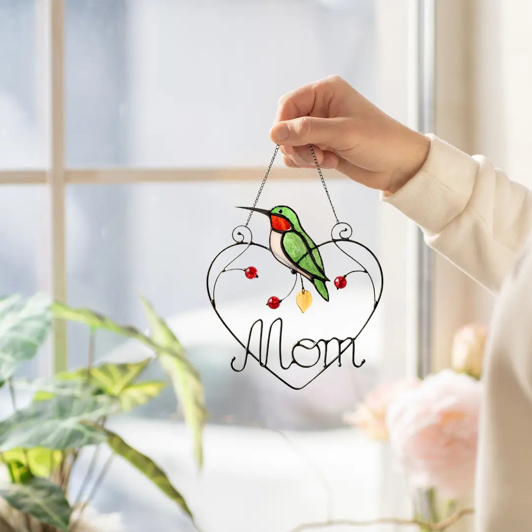 Heart-shaped stained glass ornament with a hummingbird design and 'Mom' text, held by a hand in a bright indoor setting.