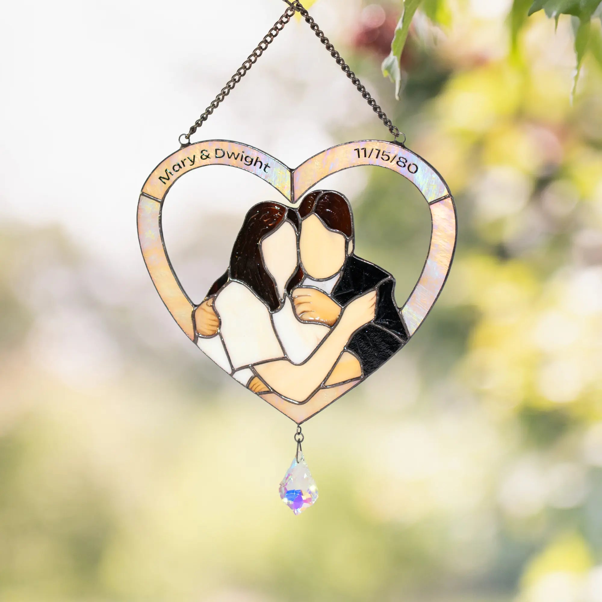 Heart-shaped stained glass suncatcher with a couple hugging, engraved names and date, against a blurred natural background.