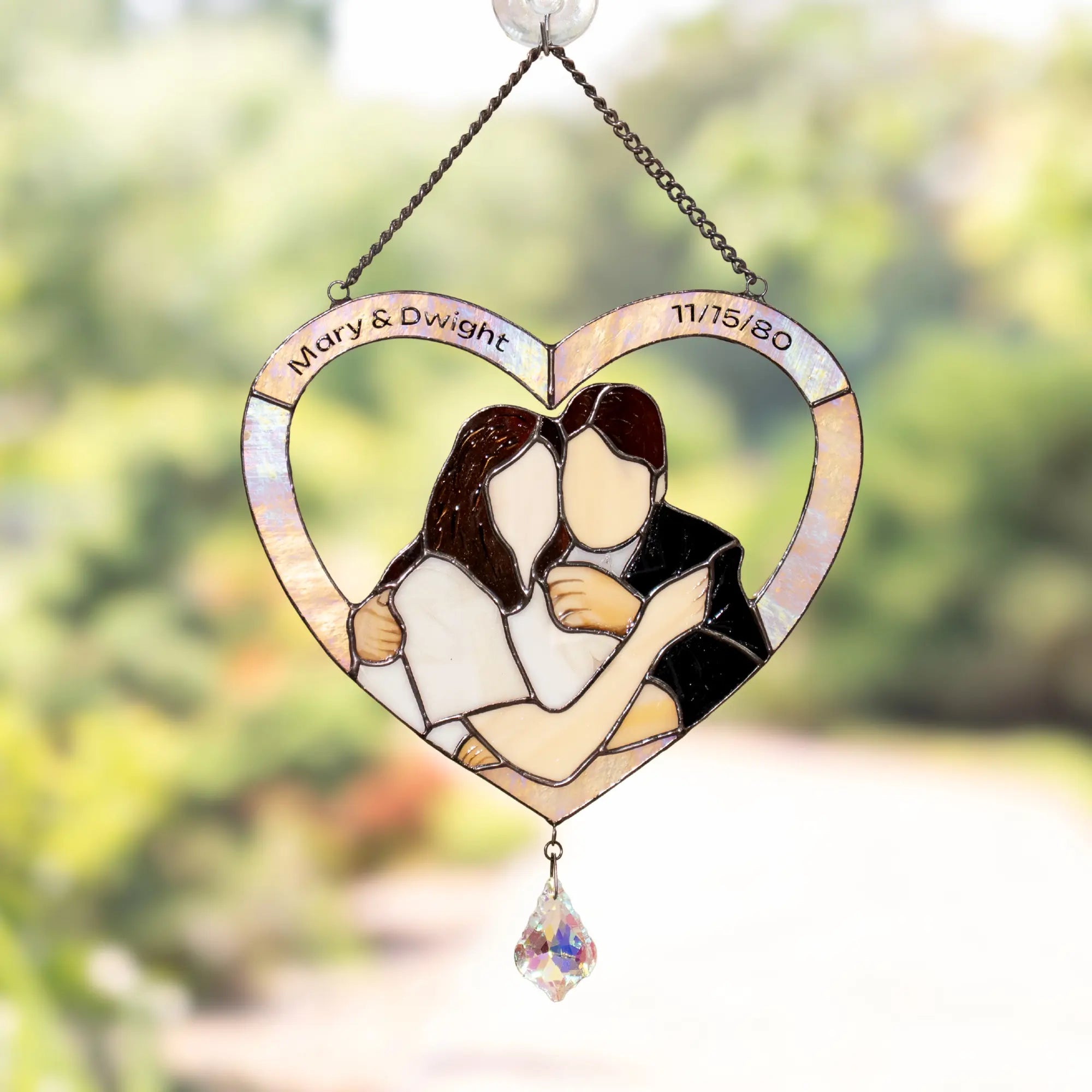 Heart-shaped stained glass panel with a couple embracing, personalized with names and date, against a blurred natural background.