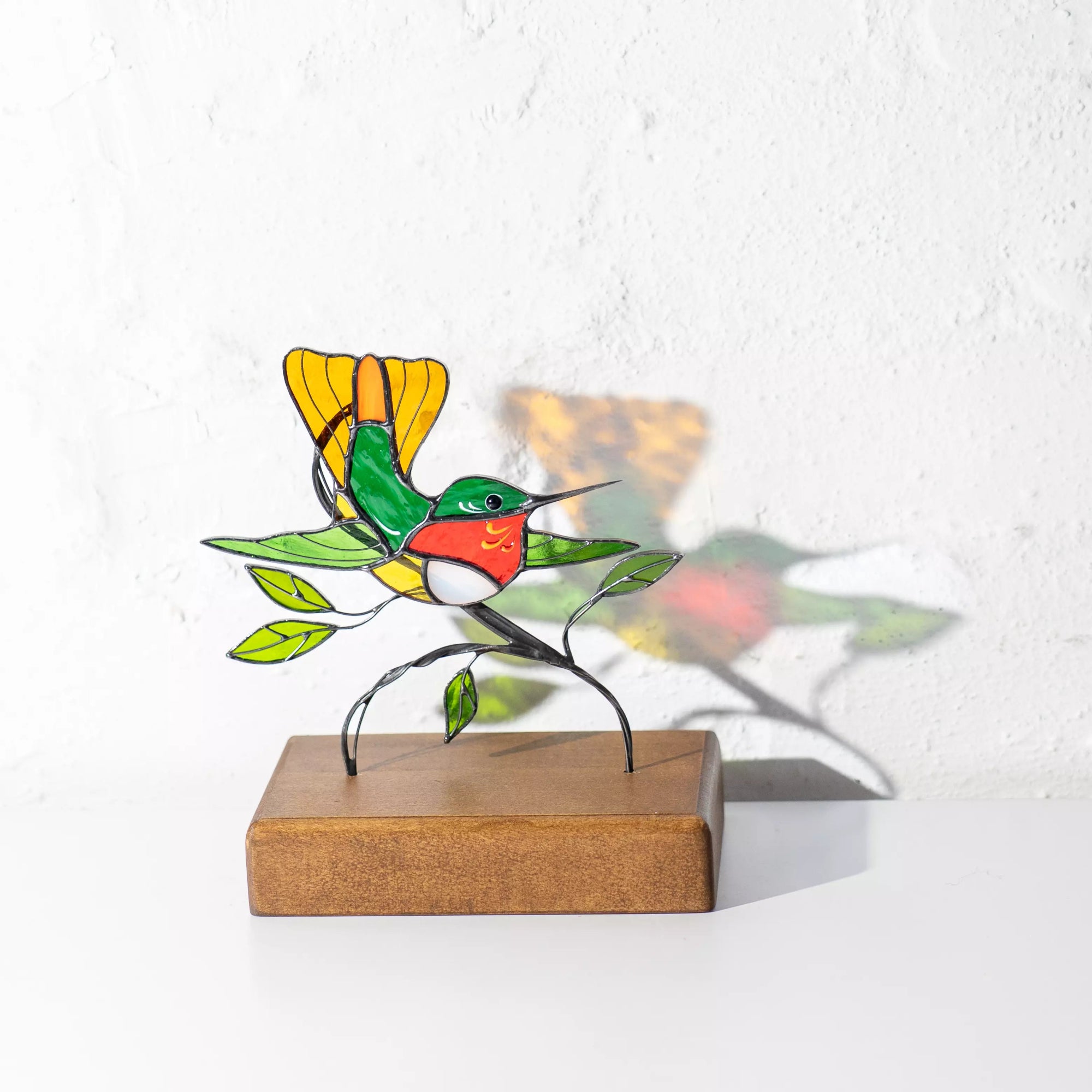Vivid stained glass hummingbird perched on branch, set against white wall with colorful reflection, creating playful shadow effect.