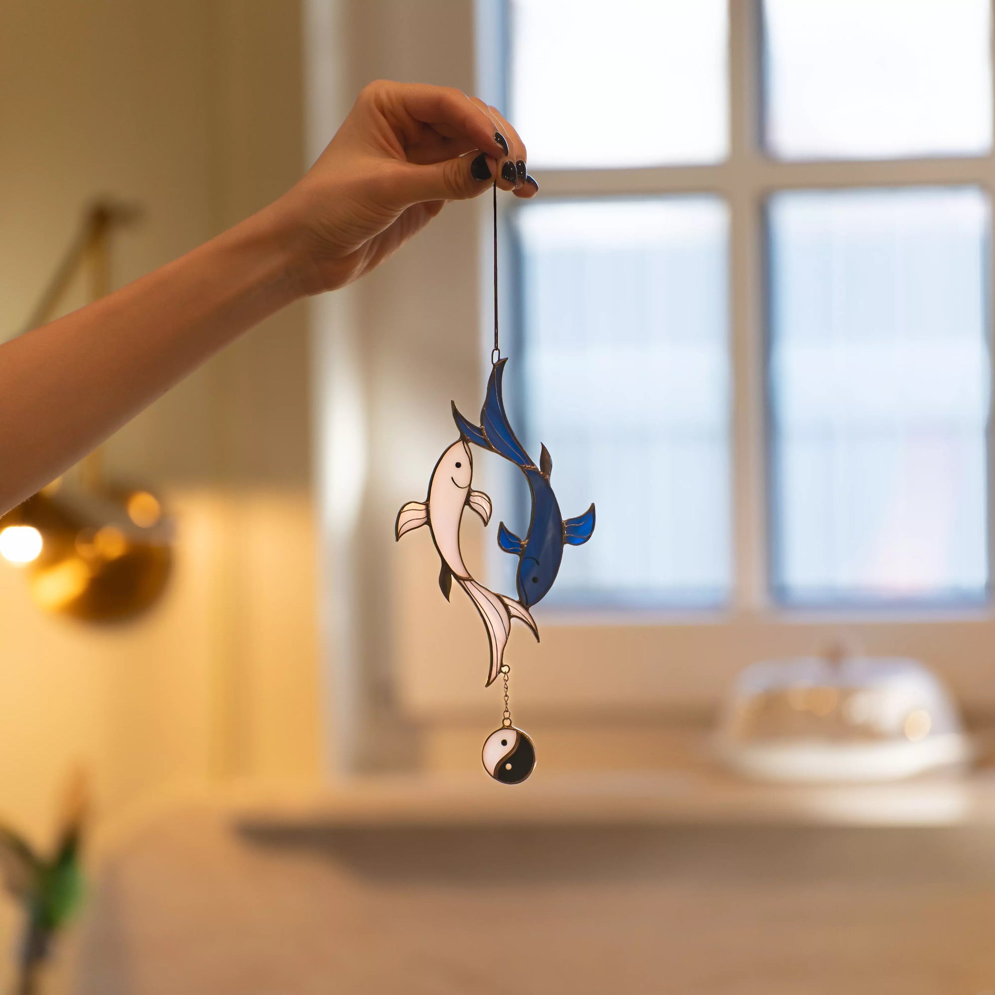 A person holding a koi fish stained glass suncatcher in a cozy, sunlit room near a window.