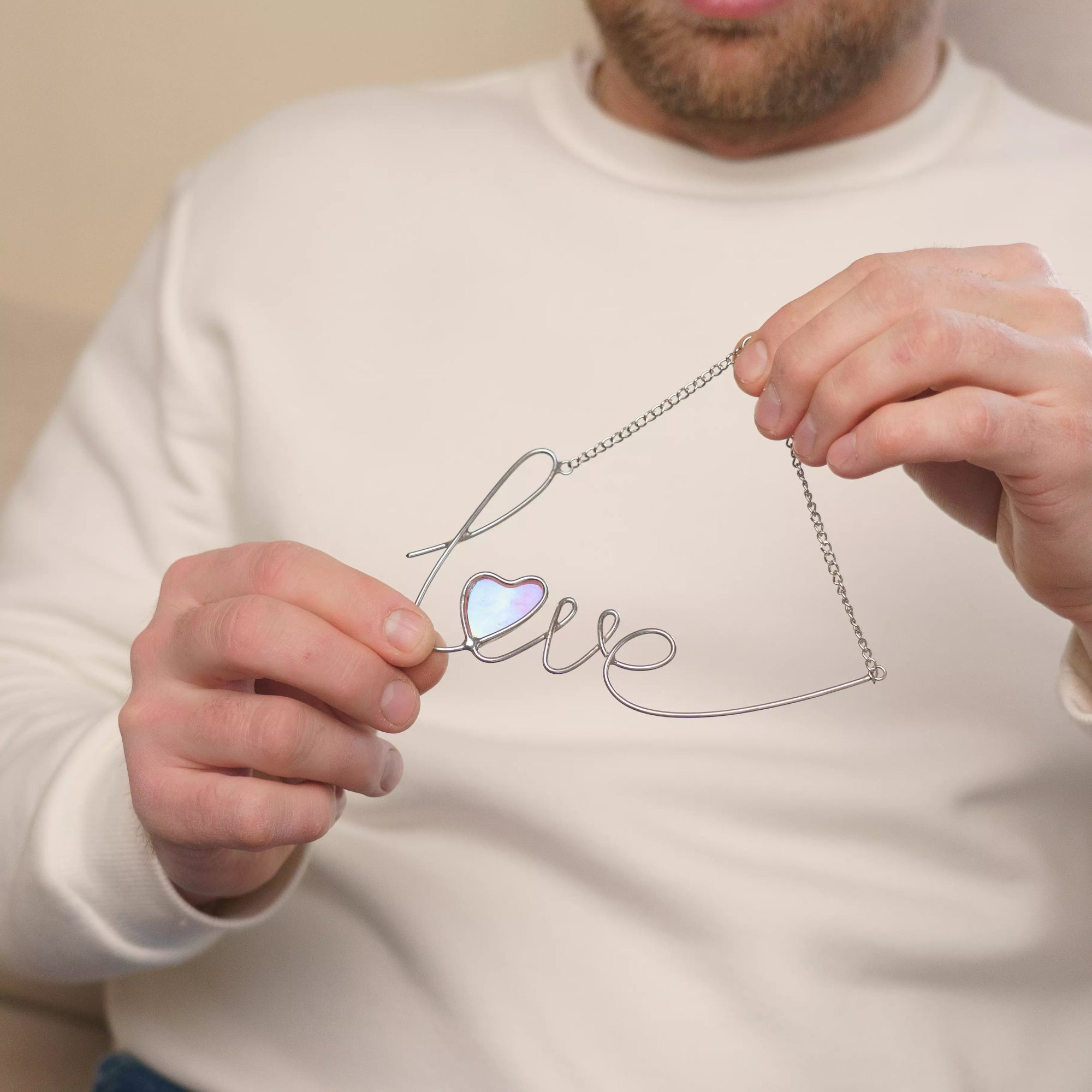 Close-up detail of the iridescent pink glass heart in a love stained glass wire sign. The handmade stained glass suncatcher reveals subtle color shifts in light, highlighting craftsmanship and the romantic character of this window decor piece.