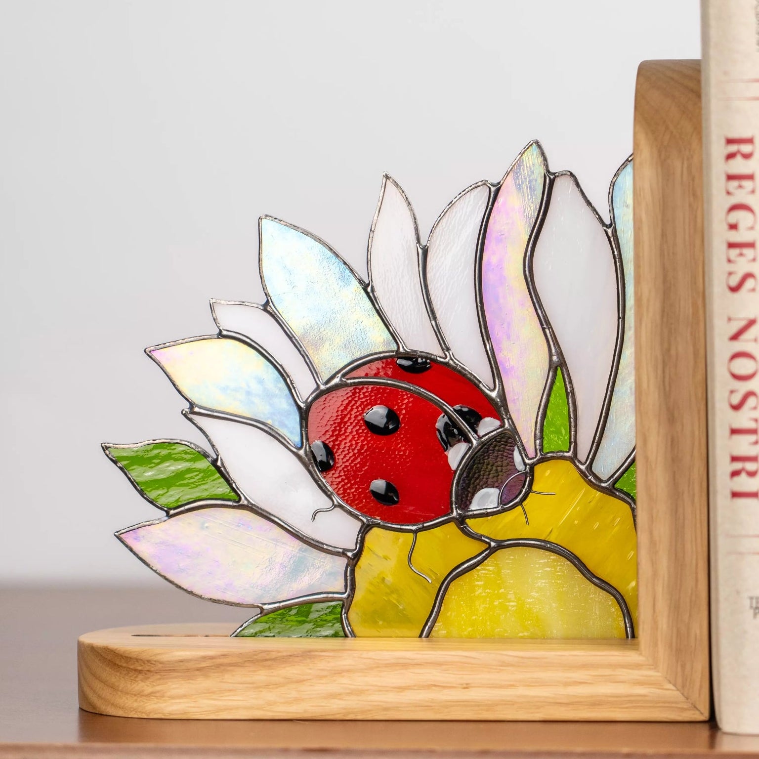 A wooden bookend with a stained glass ladybug and daisy design stands on a shelf. The iridescent white petals and red ladybug look elegant against the books, enhancing the shelf aesthetic