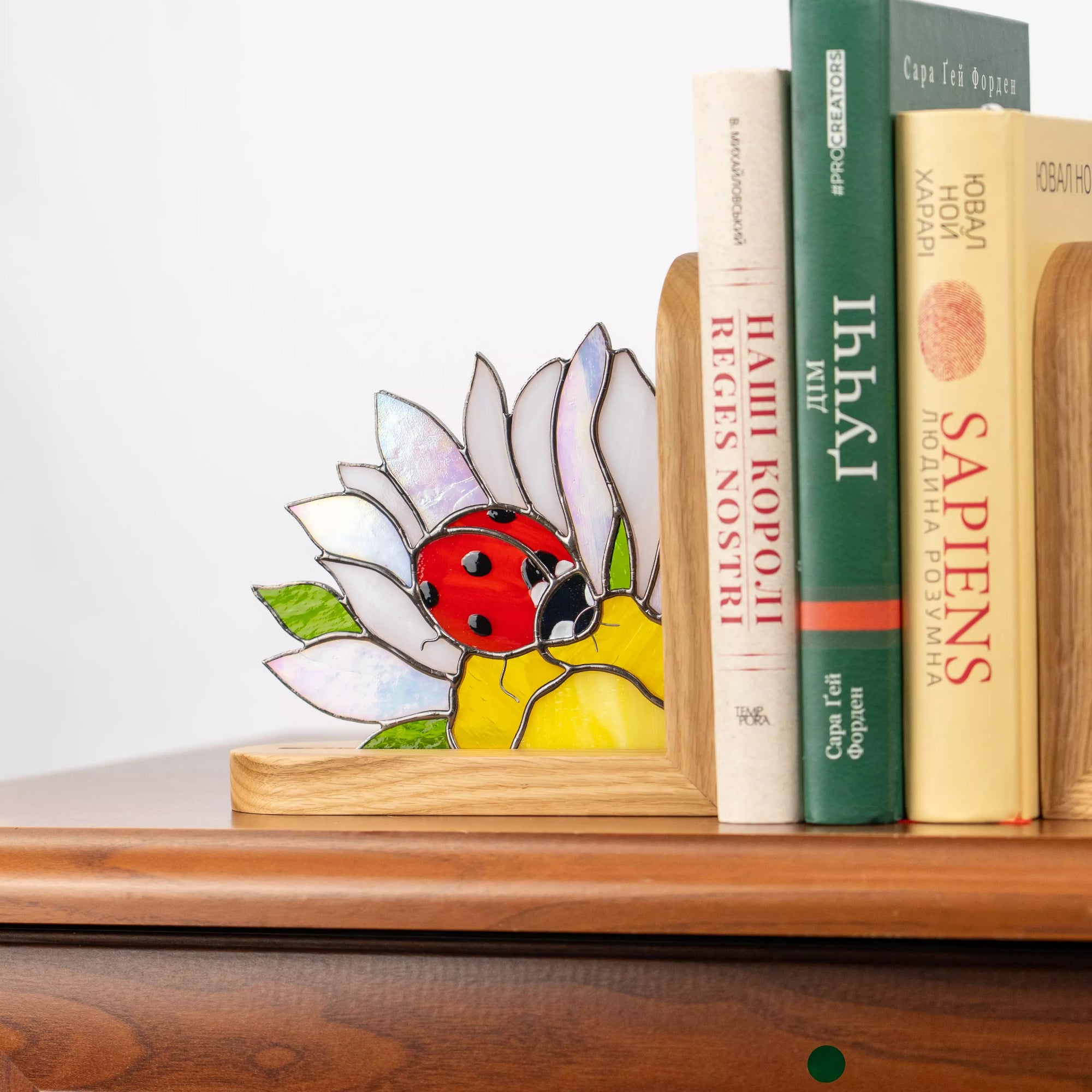 A wooden bookend with a stained glass ladybug and daisy on a shelf. The iridescent petals and red beetle add a pop of color, perfectly organizing and beautifying your home library.