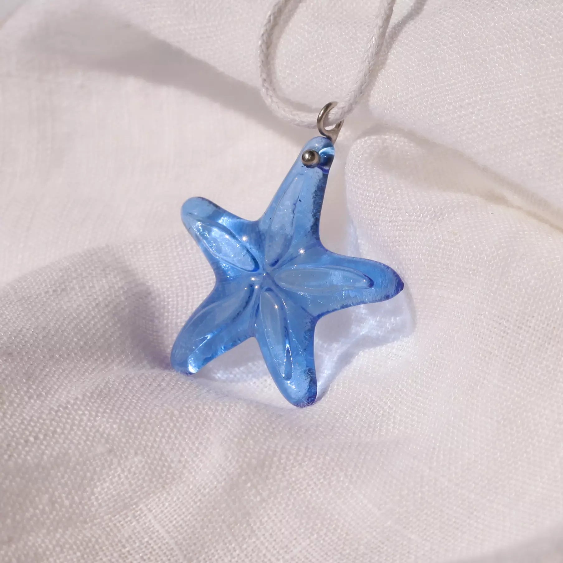 Light sapphire starfish glass pendant on white cord, displayed on a textured white linen fabric.